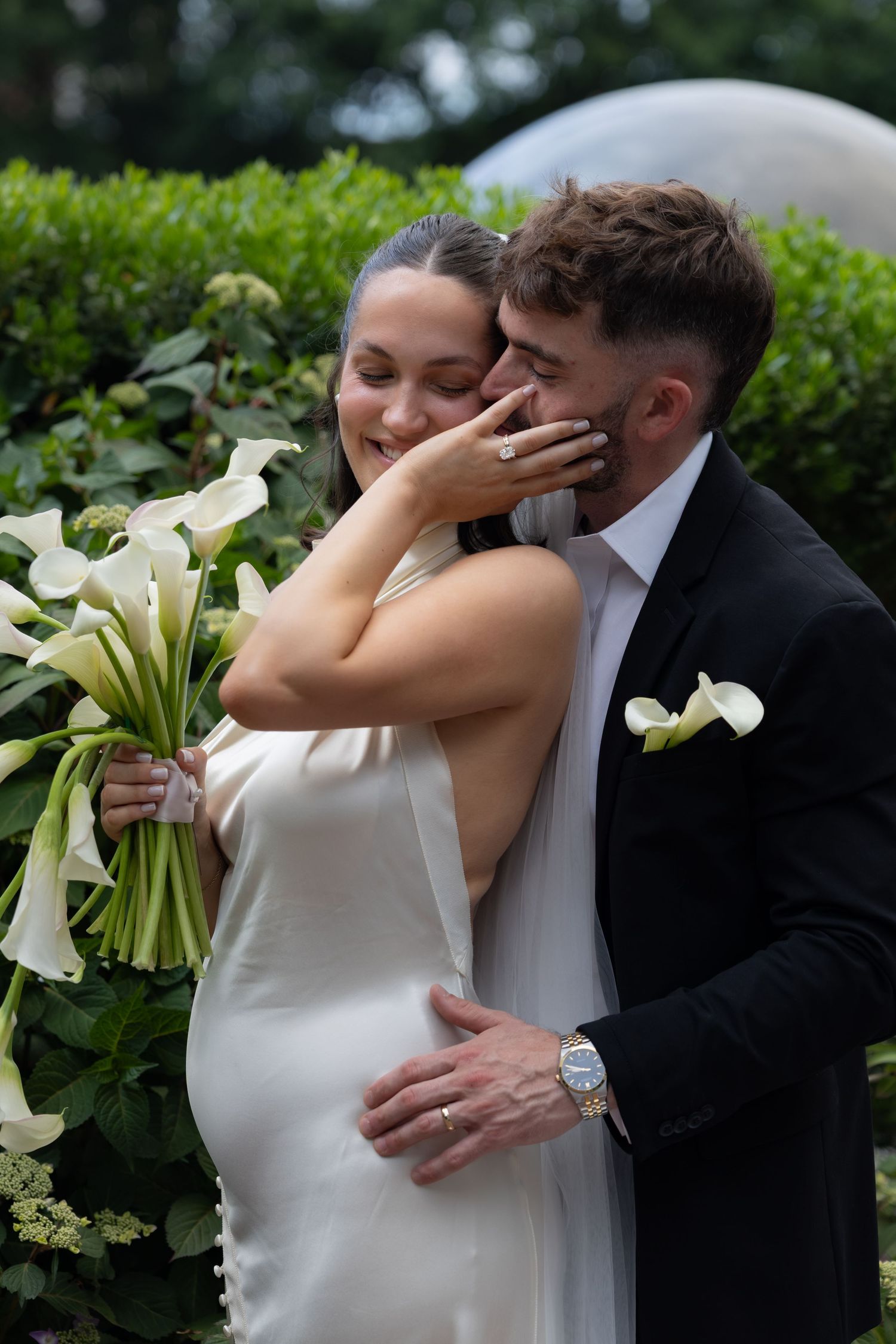 A bride holding white calla lilies embraces her groom in a black suit outdoors, sharing a tender wedding moment.