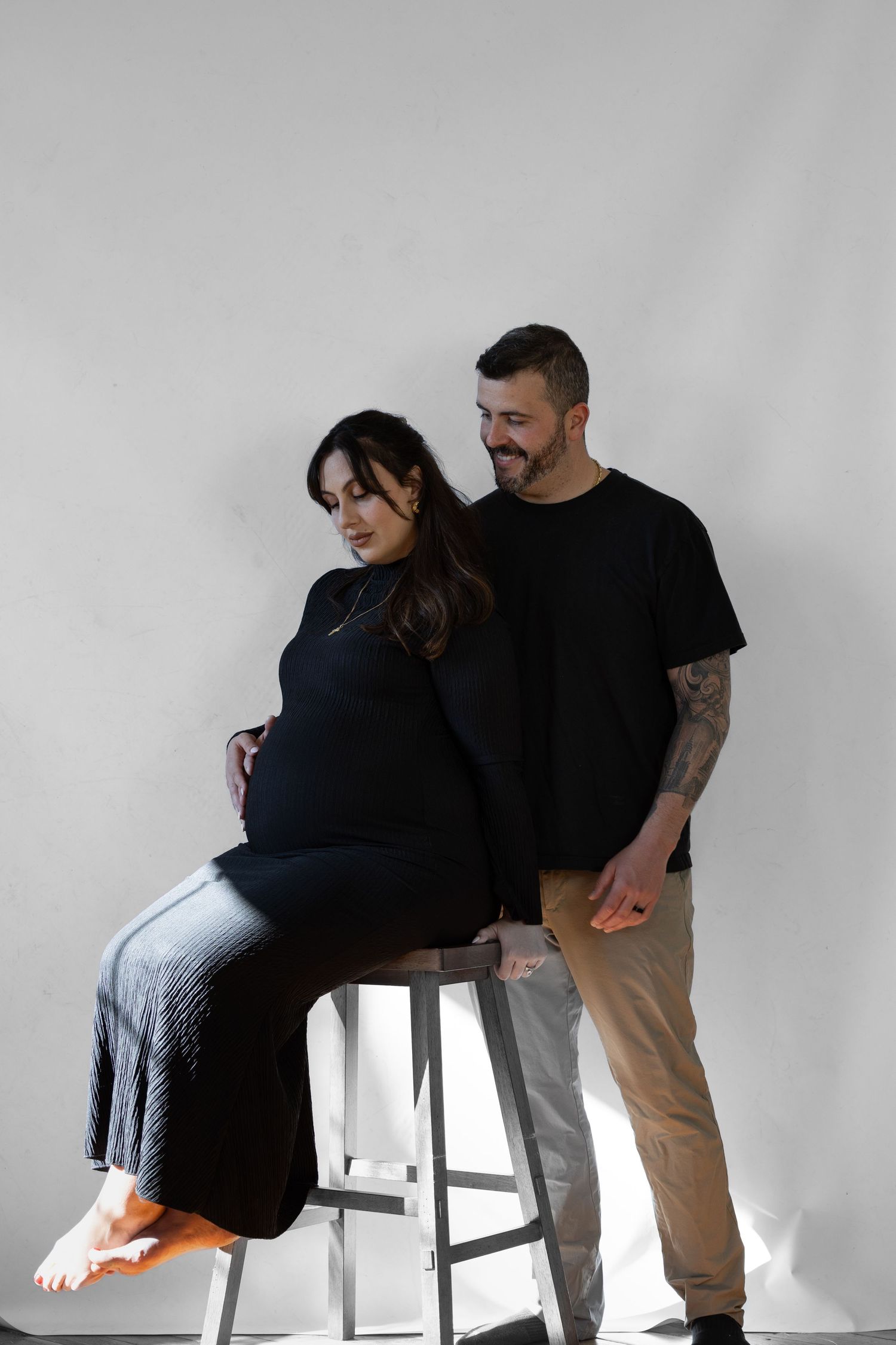 Smiling couple expecting a baby, woman in black dress seated on stool with man standing behind.