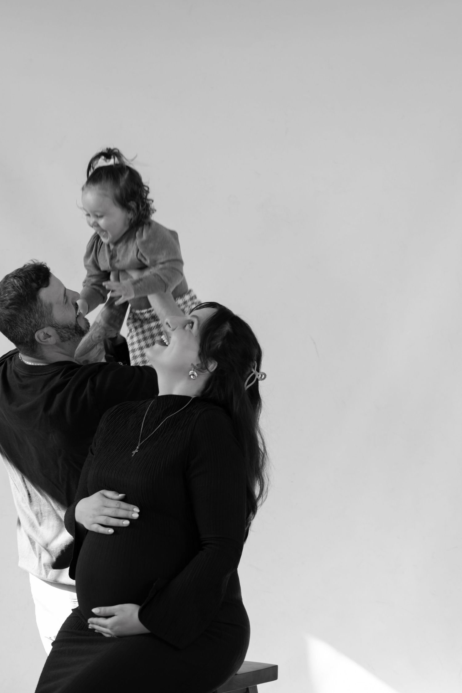 Black and white photo of pregnant woman and man lifting toddler girl, happy family maternity portrait studio session.