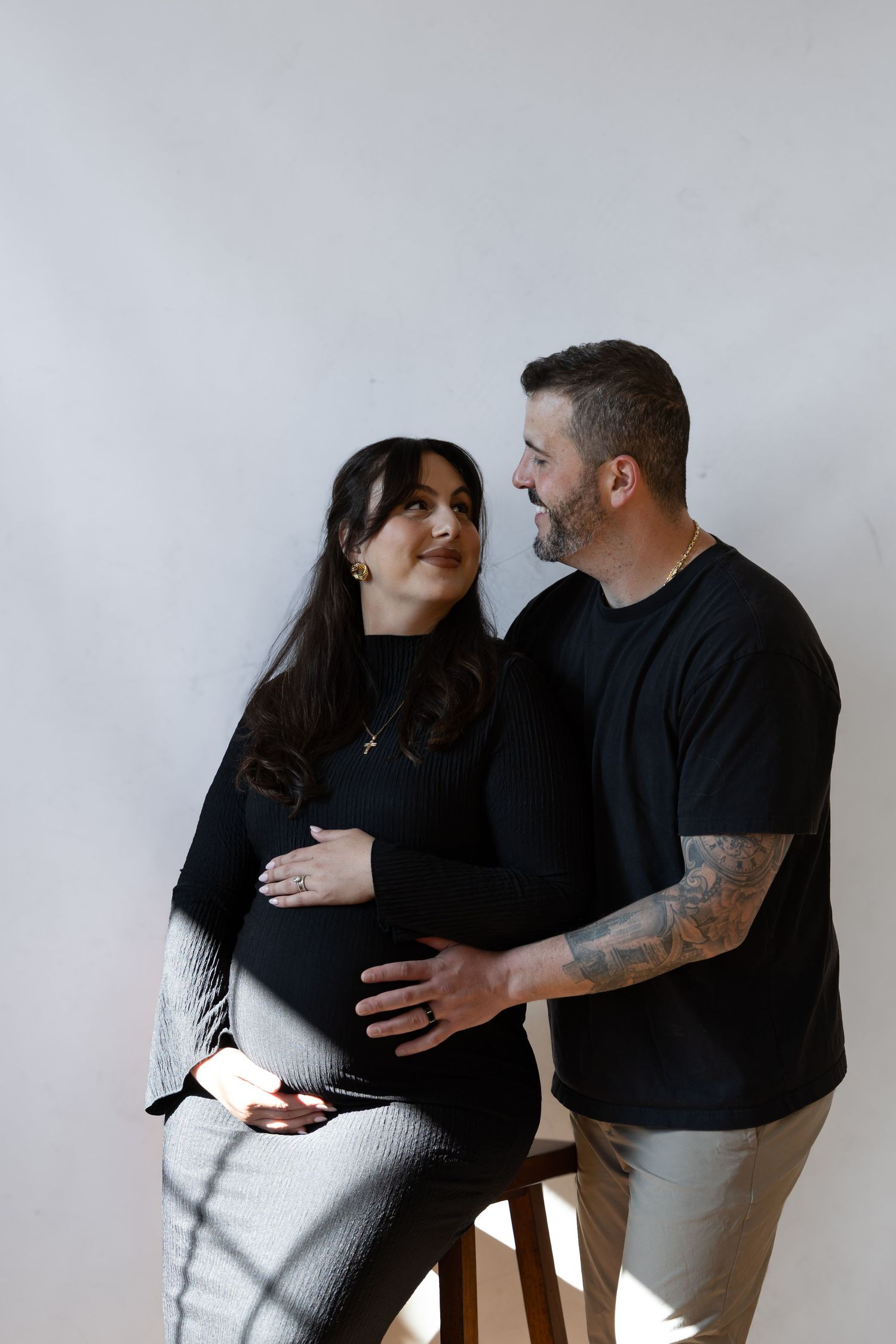 Pregnant woman in black outfit sits on stool as tattooed man in black tee tenderly holds her baby bump.
