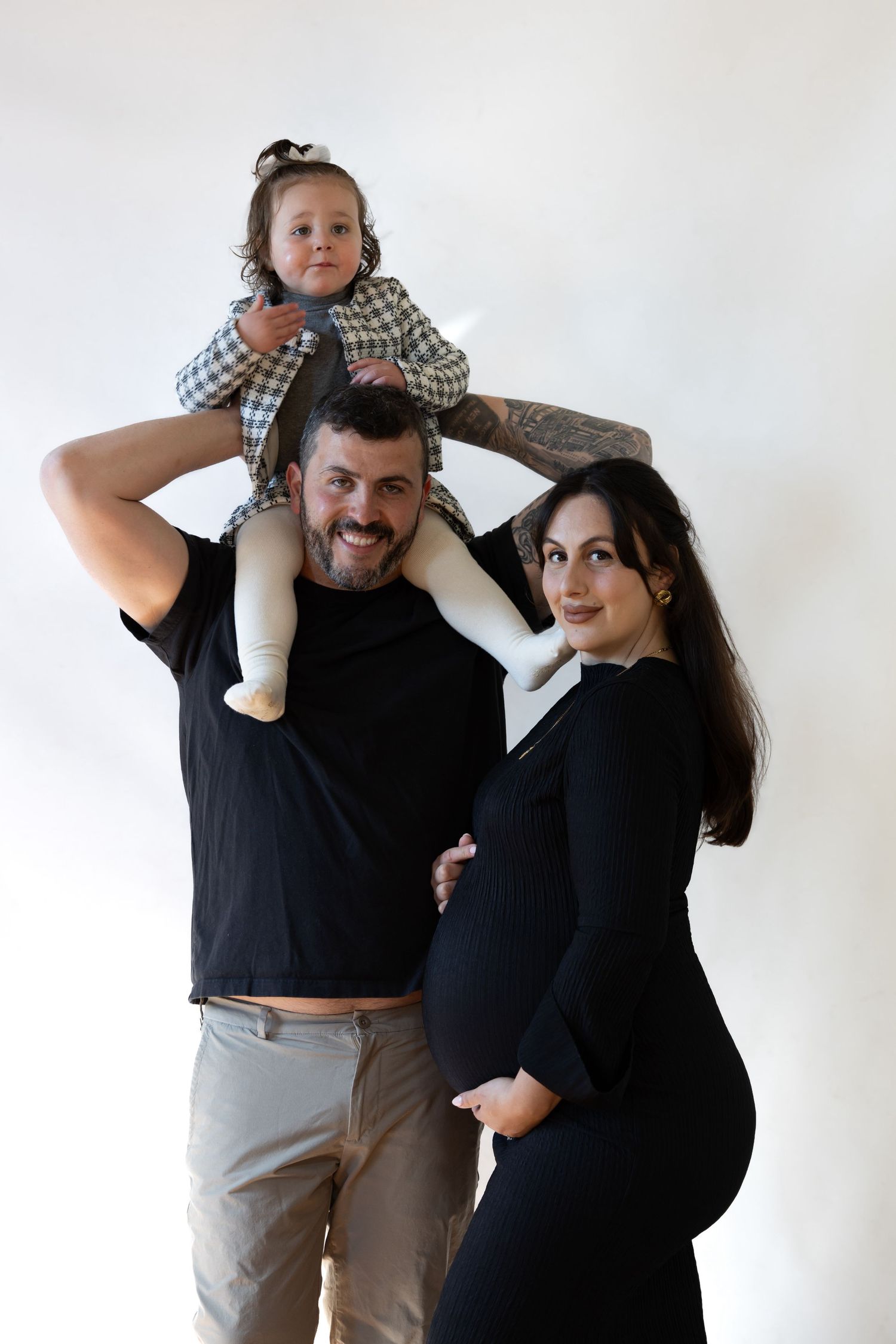 Smiling family portrait with bearded man, toddler on shoulders, and pregnant woman in black dress.