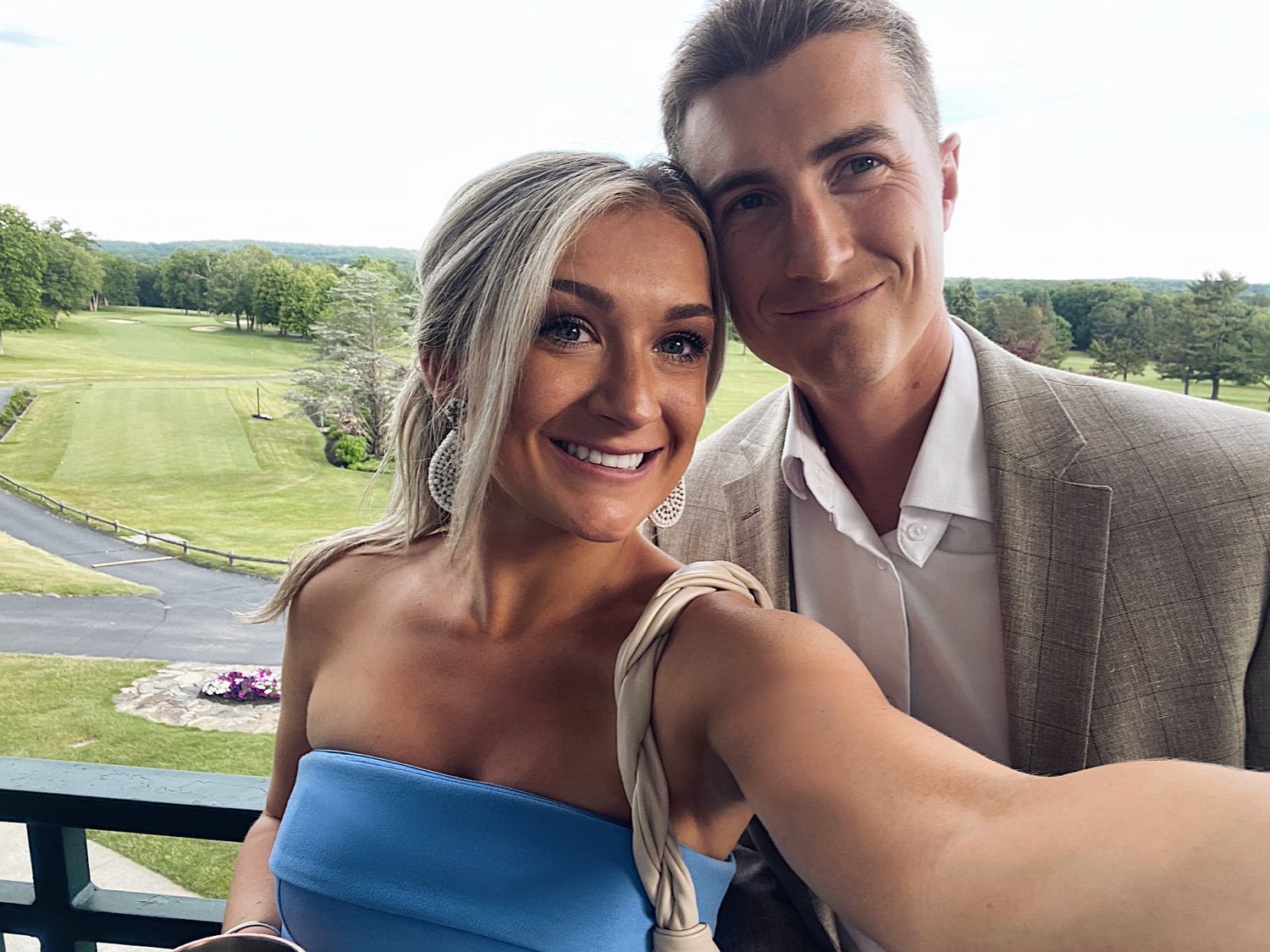 A couple takes a selfie together on a balcony overlooking a scenic golf course on a sunny day.