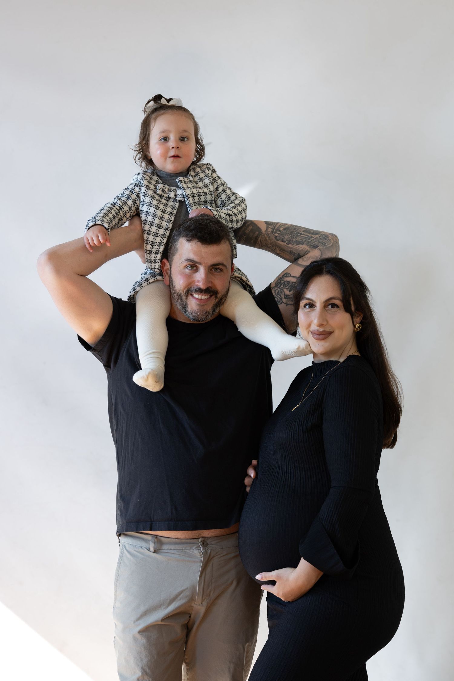 Happy family maternity photo with pregnant woman, man, and toddler on shoulders against white background.