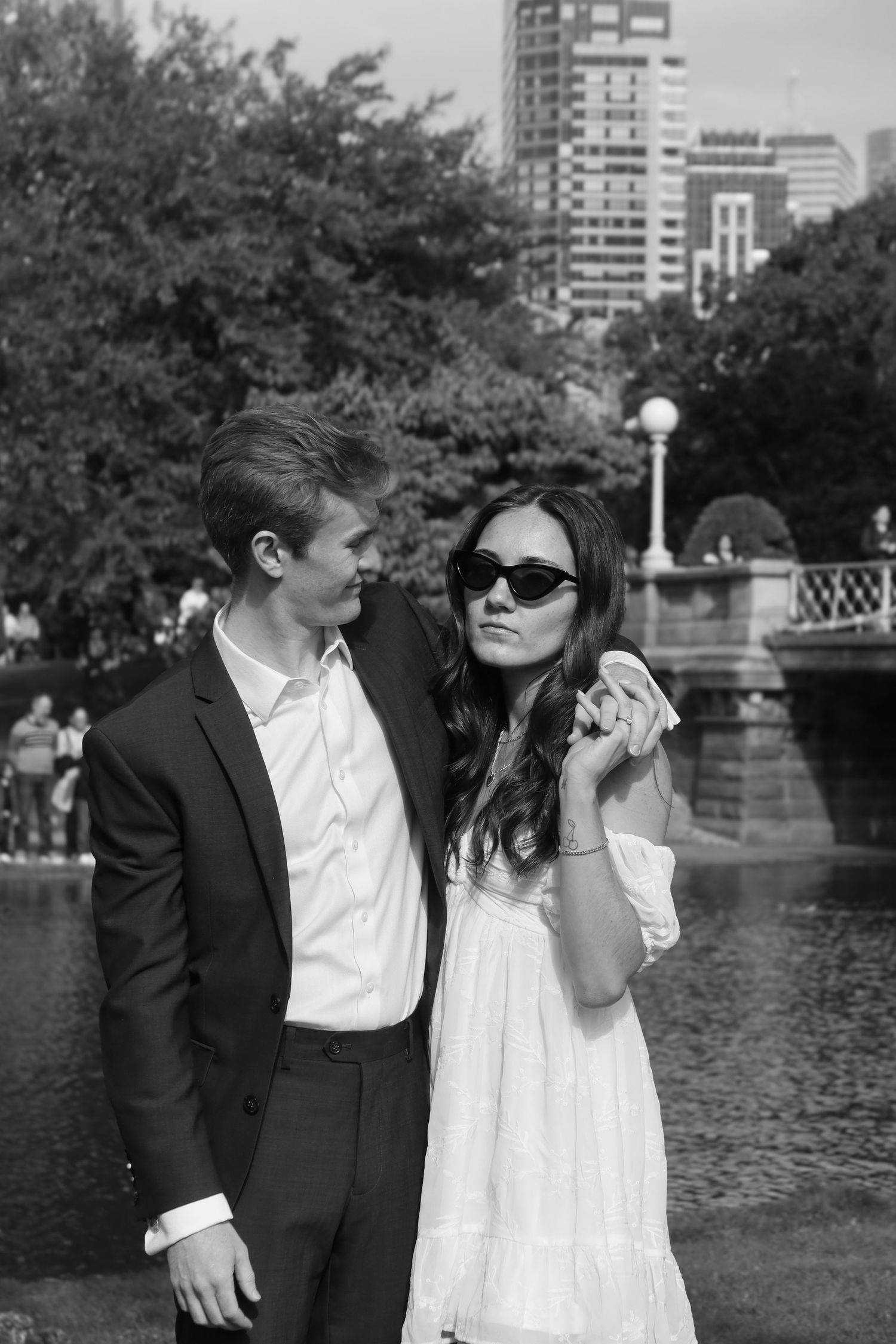 A couple shares a playful moment by a pond in a park with city buildings in the background.