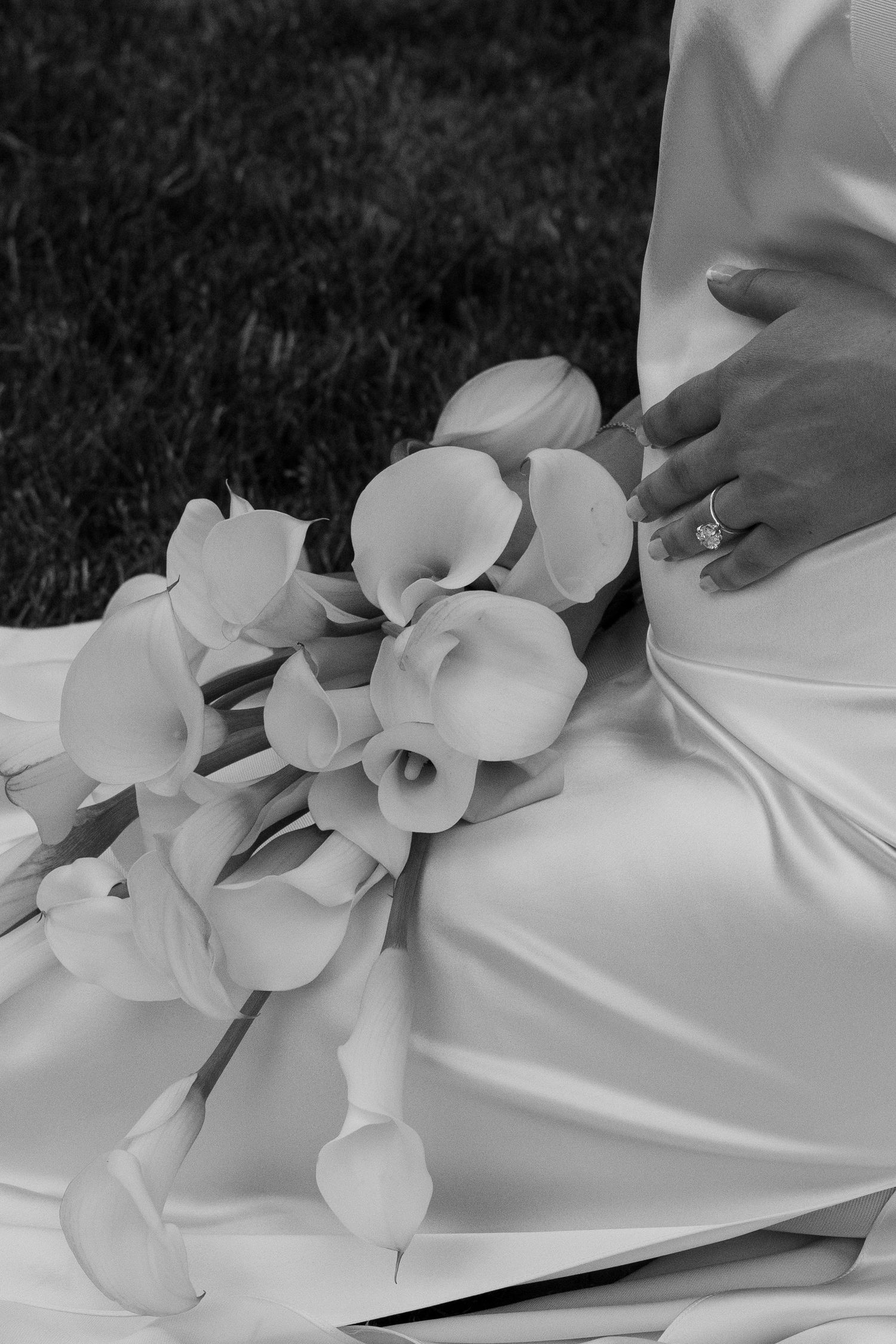 Black and white photo of a bride in a white gown holding a bouquet of calla lilies, with a diamond ring visible.