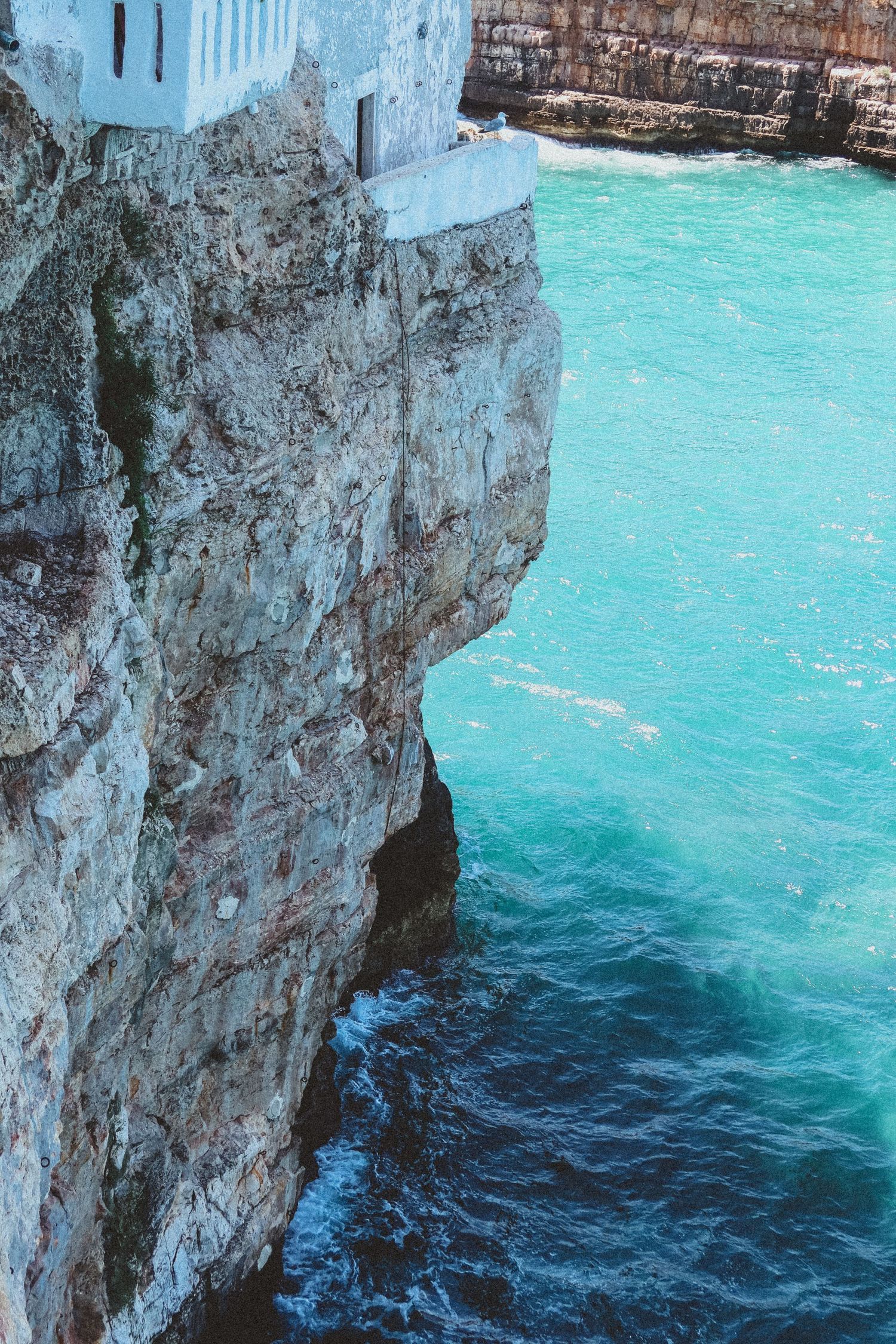 Stunning turquoise Mediterranean waters crash against white limestone cliffs near coastal buildings.