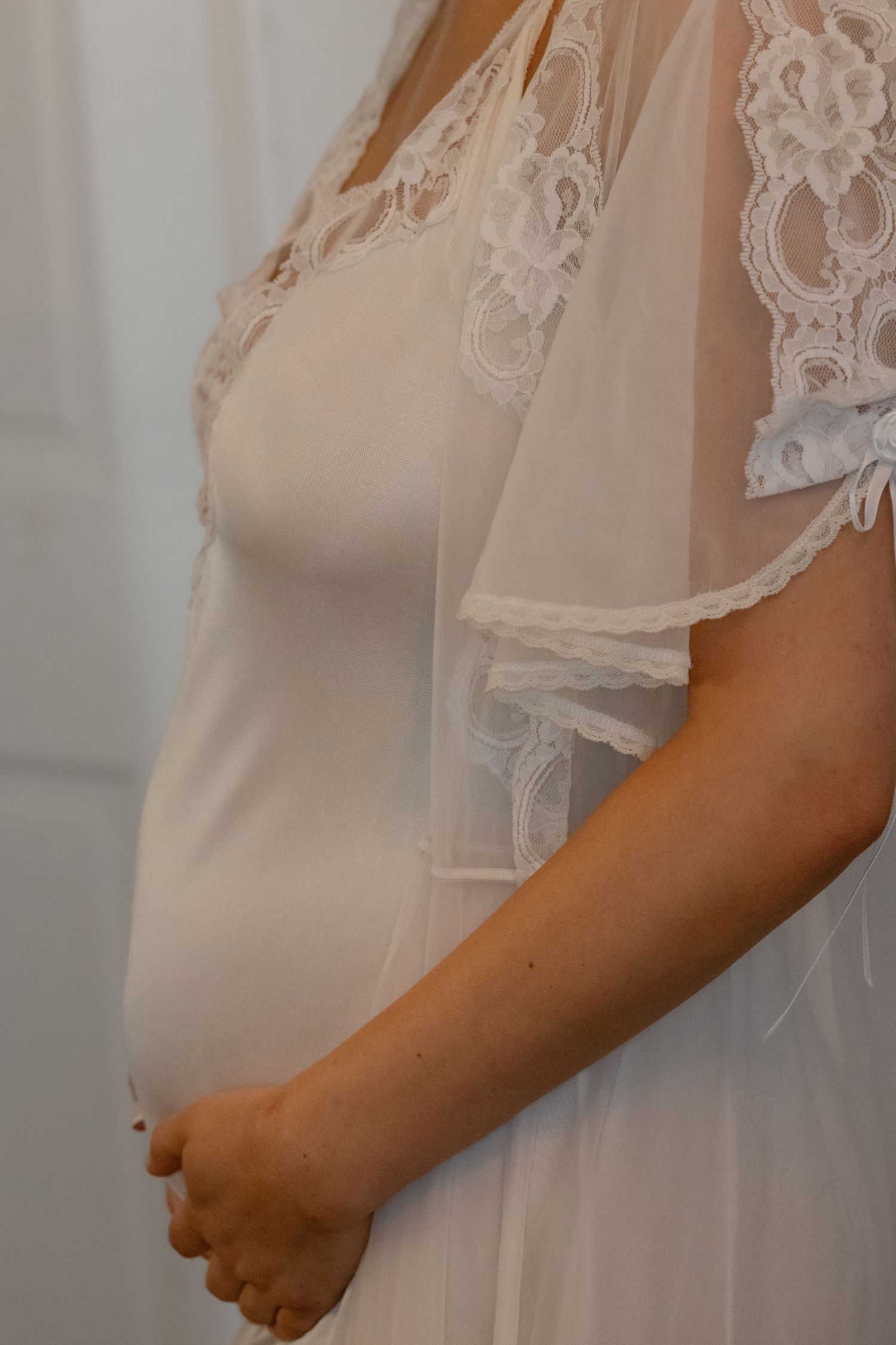 Close-up side profile of pregnant bride in white dress with lace cape veil showing baby bump detail.