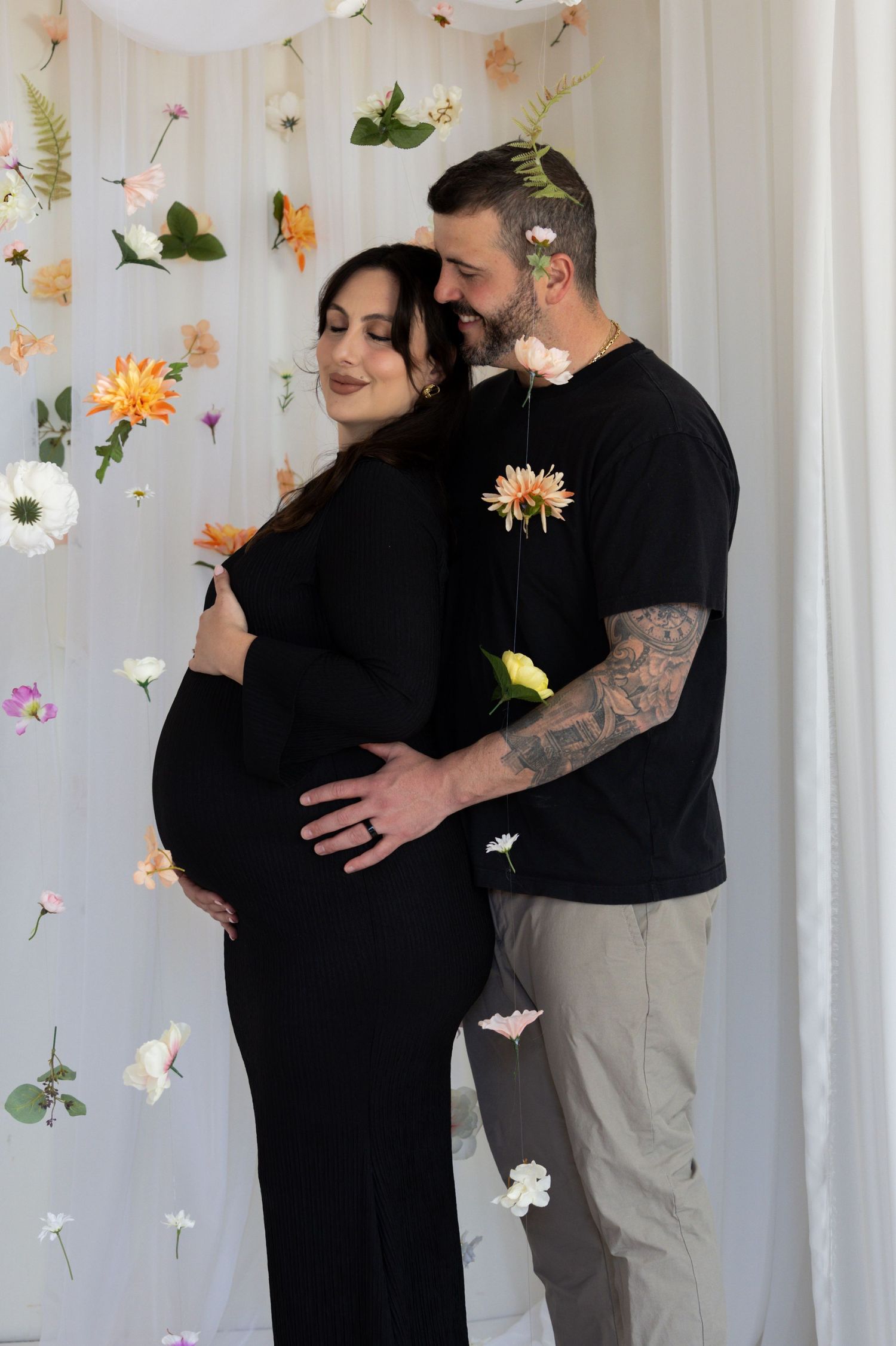 Couple poses for maternity photo shoot with floral backdrop, woman in black dress cradling baby bump.