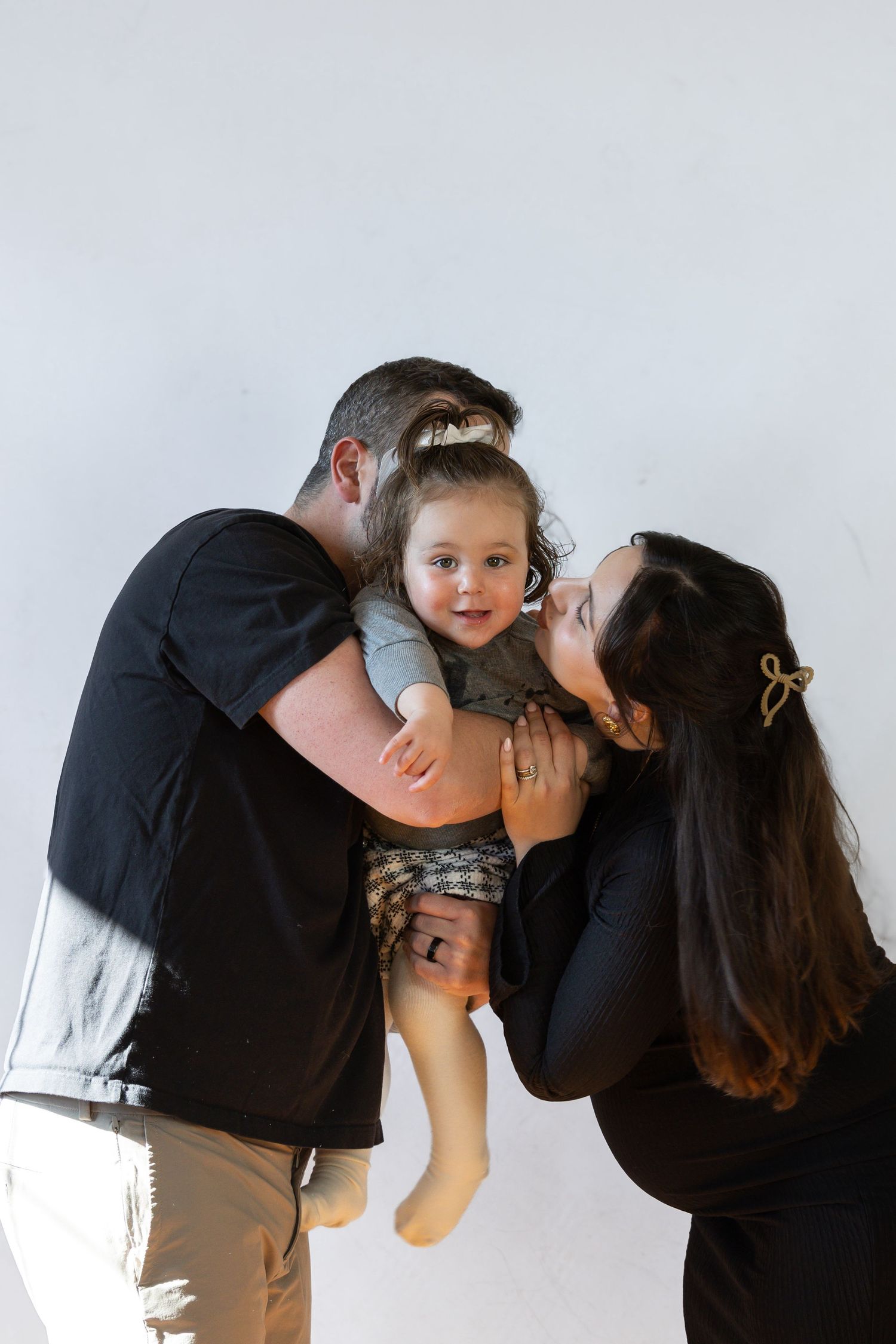 Two parents in black shirts kiss their smiling baby on the cheeks against a white background.