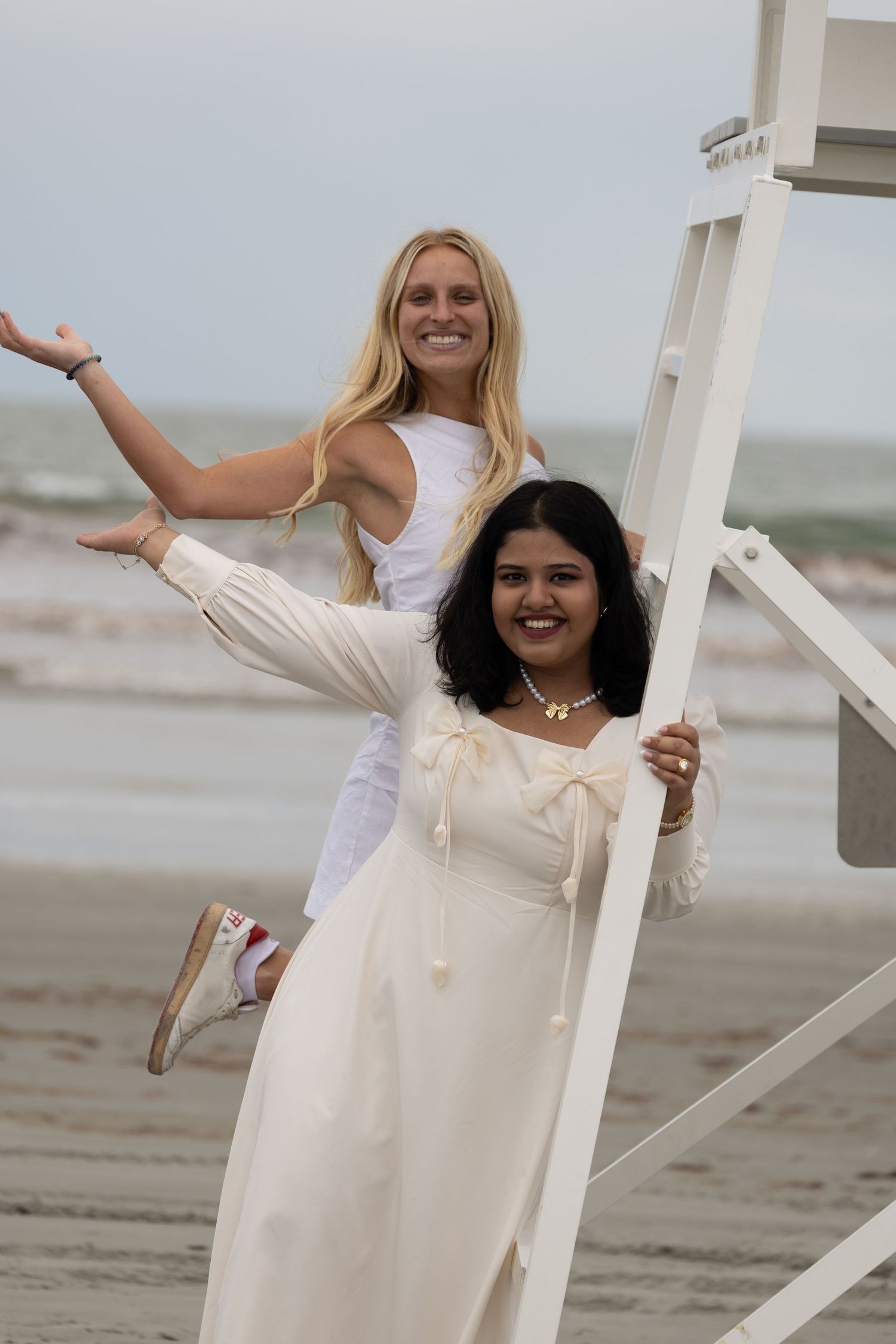 Two smiling women in white dresses pose playfully by a lifeguard stand on a cloudy beach.
