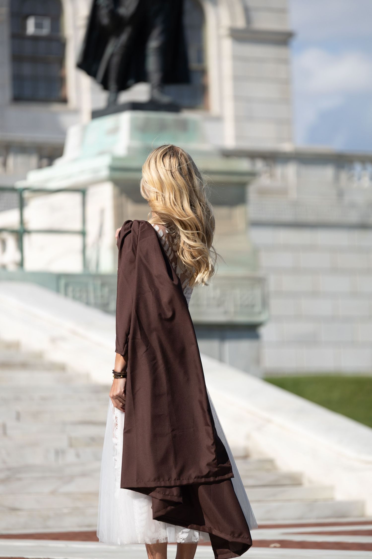 Blonde woman in brown graduation gown and white dress stands before a monument on marble steps outdoors.