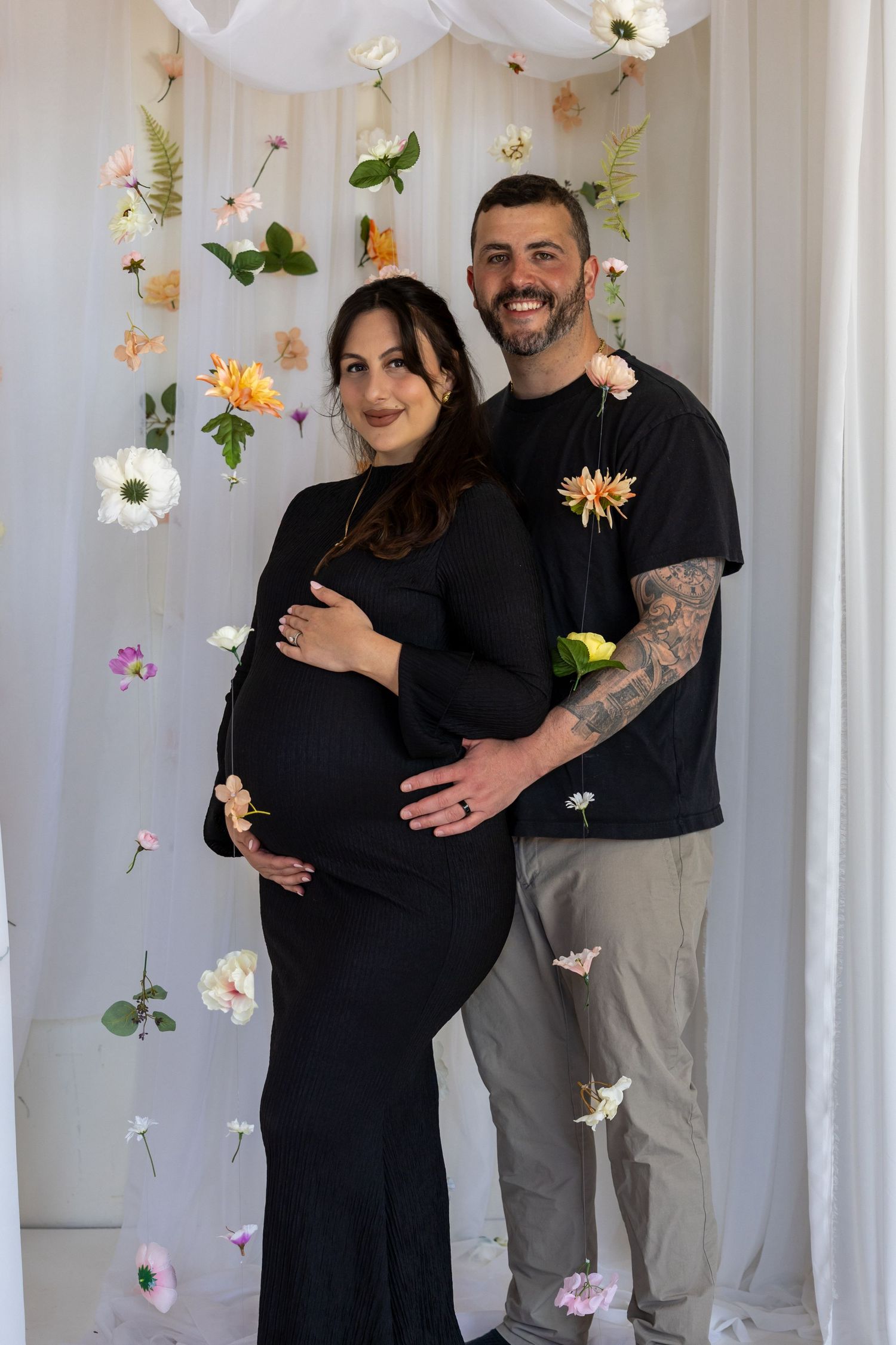 Smiling couple posing for maternity photo in black outfits against floral backdrop with hanging flowers.