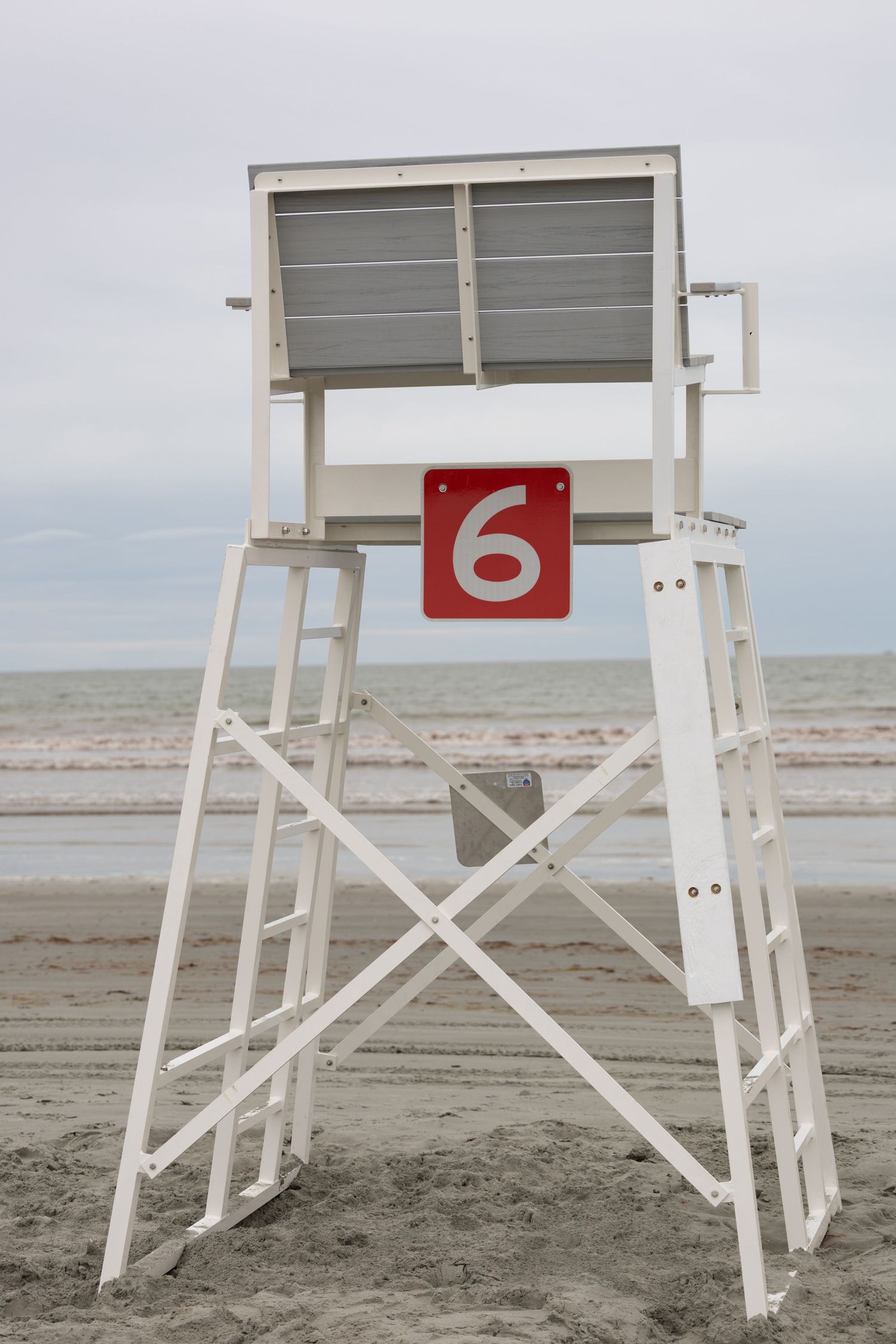 White lifeguard tower number 6 on an empty sandy beach with overcast skies and ocean waves in the background.