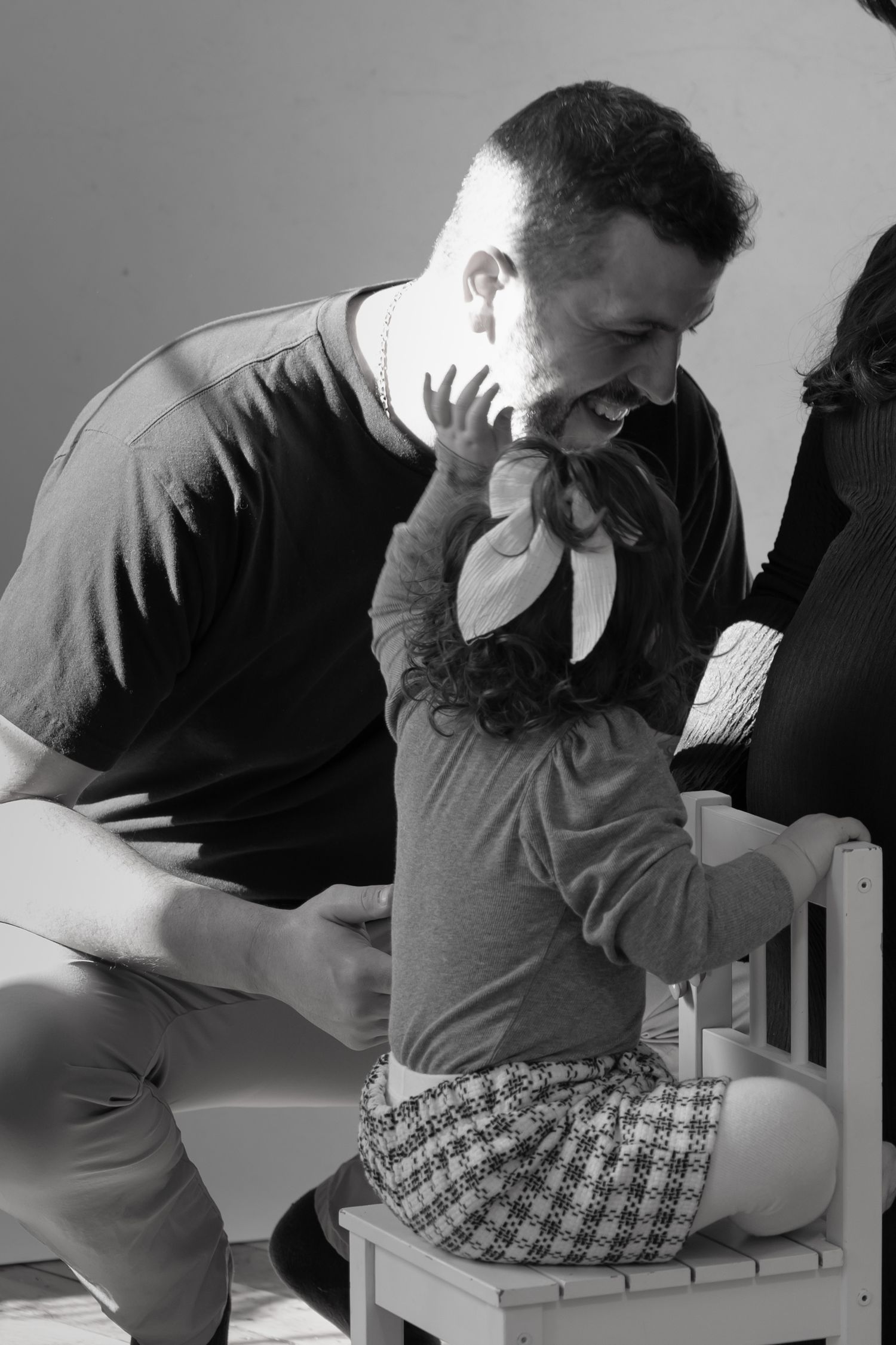 Father laughing and playing with young curly-haired girl sitting on small white chair in black and white photo.