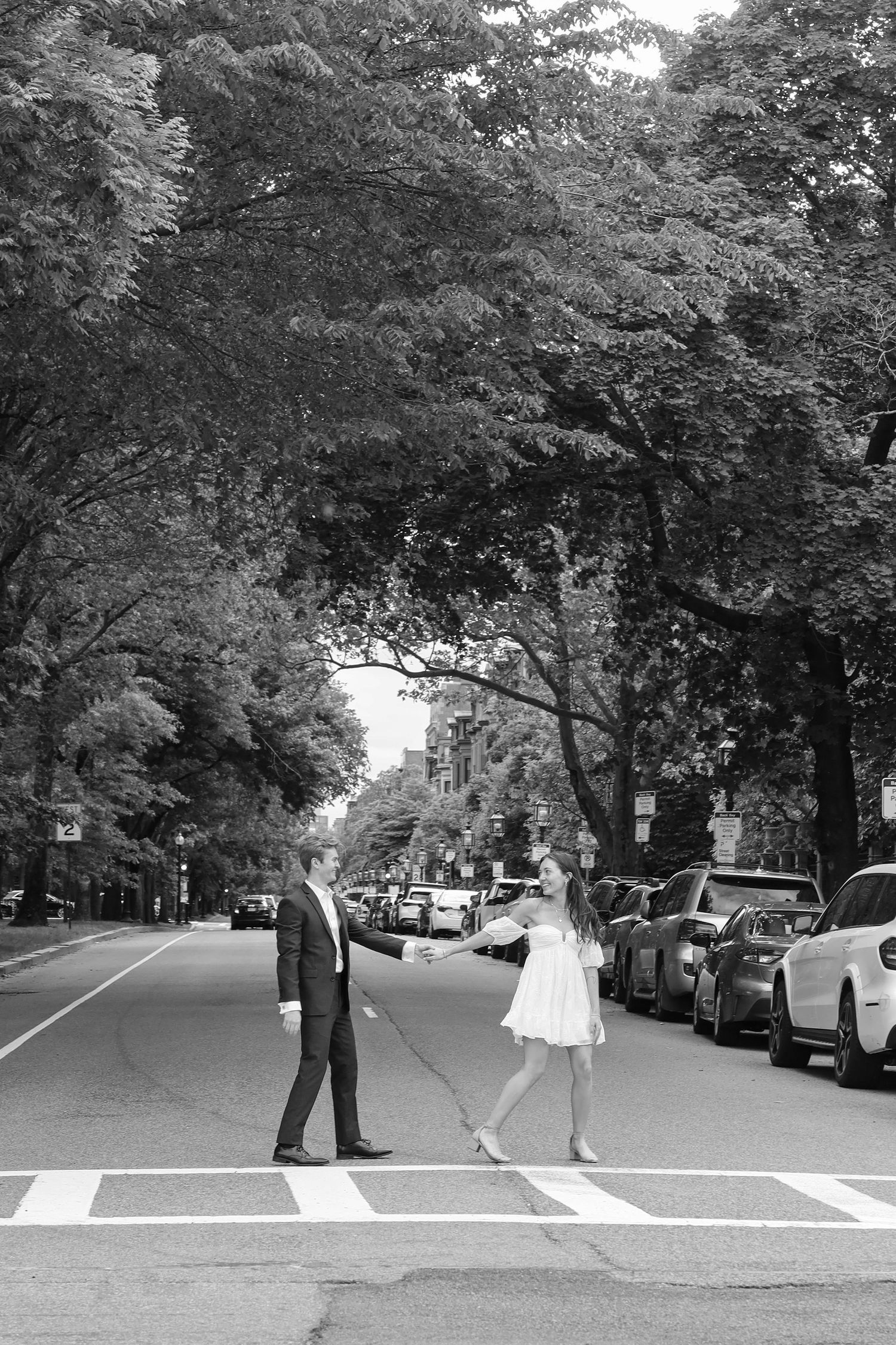 A couple poses on a tree-lined urban street with parked cars in black and white.