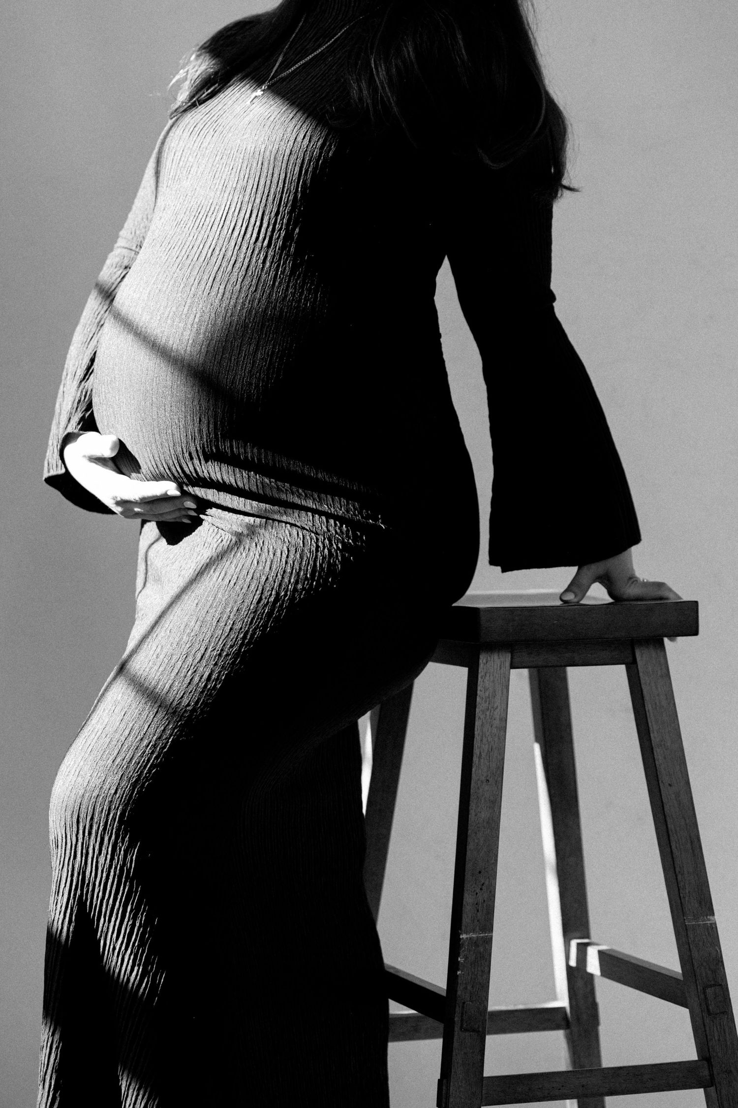 Black and white maternity photo of pregnant woman in ribbed dress leaning on wooden stool, showcasing baby bump.
