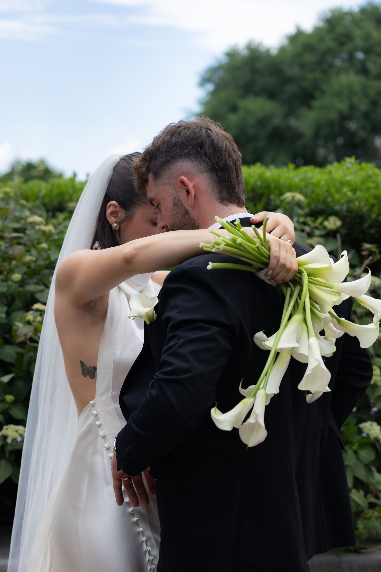 Bride and groom sharing a romantic kiss, bride holding white calla lily bouquet, wearing veil and backless gown outdoors.