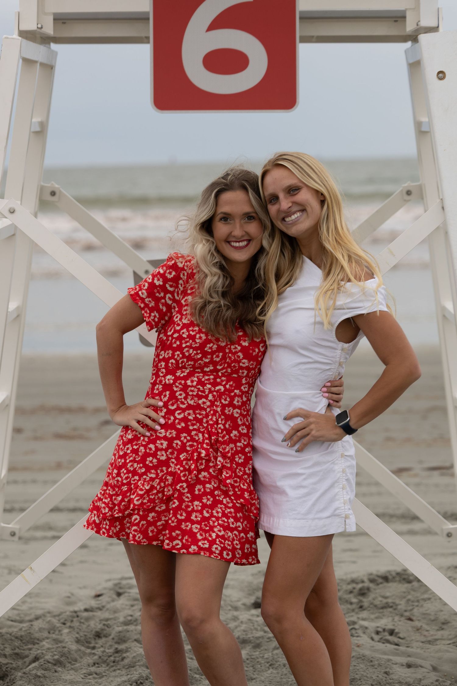 Two smiling women pose in front of lifeguard stand 6 on a sandy beach, wearing red floral and white outfits.