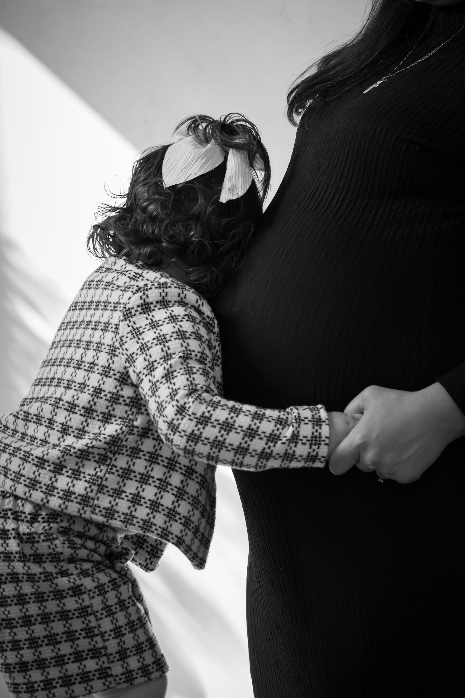 Black and white photo of young child in plaid jacket embracing pregnant mothers belly tenderly.