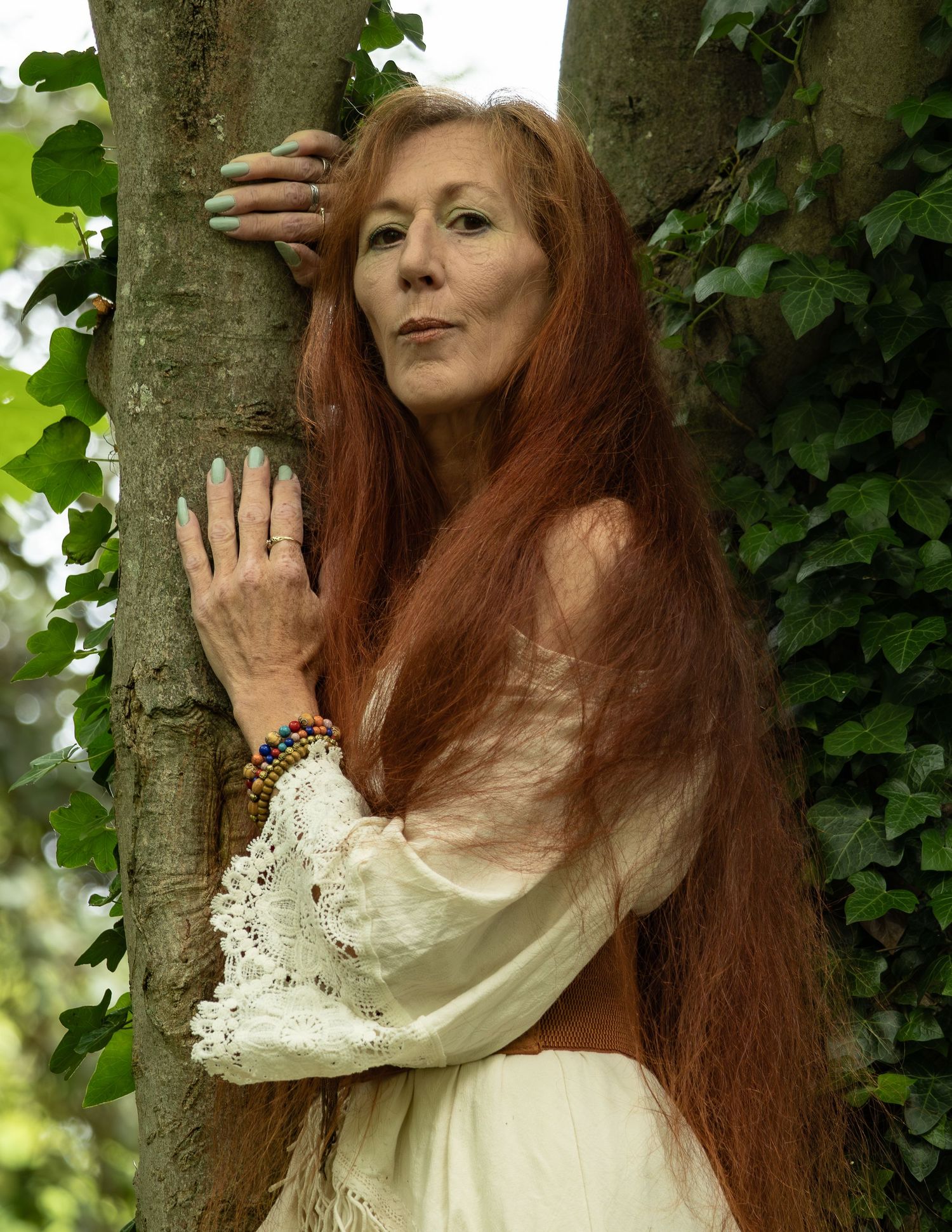 Older woman with long red hair in white lace blouse leaning against a tree in lush green garden.