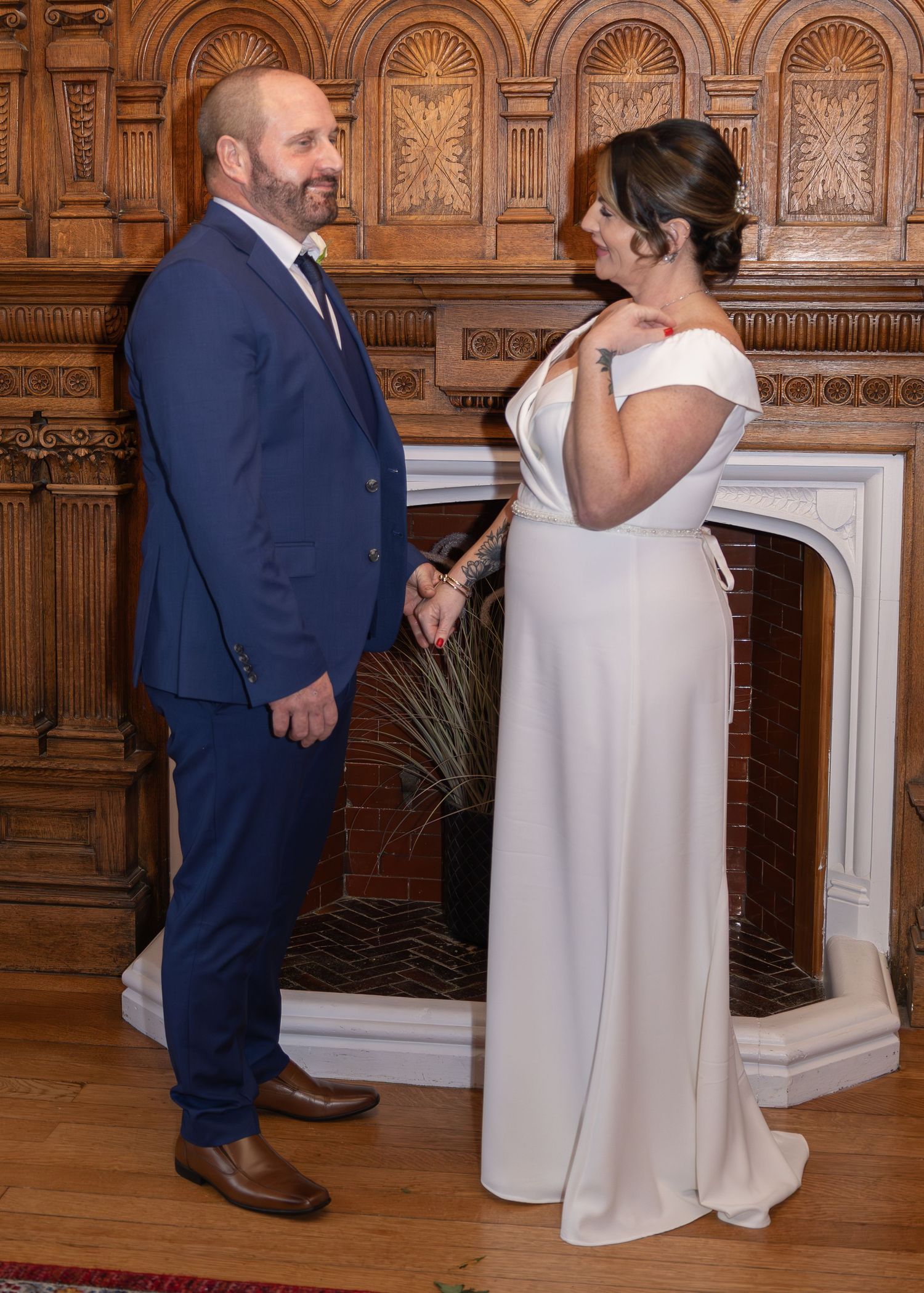 Couple in wedding attire standing by ornate fireplace, groom in navy suit, bride in white off-shoulder gown.