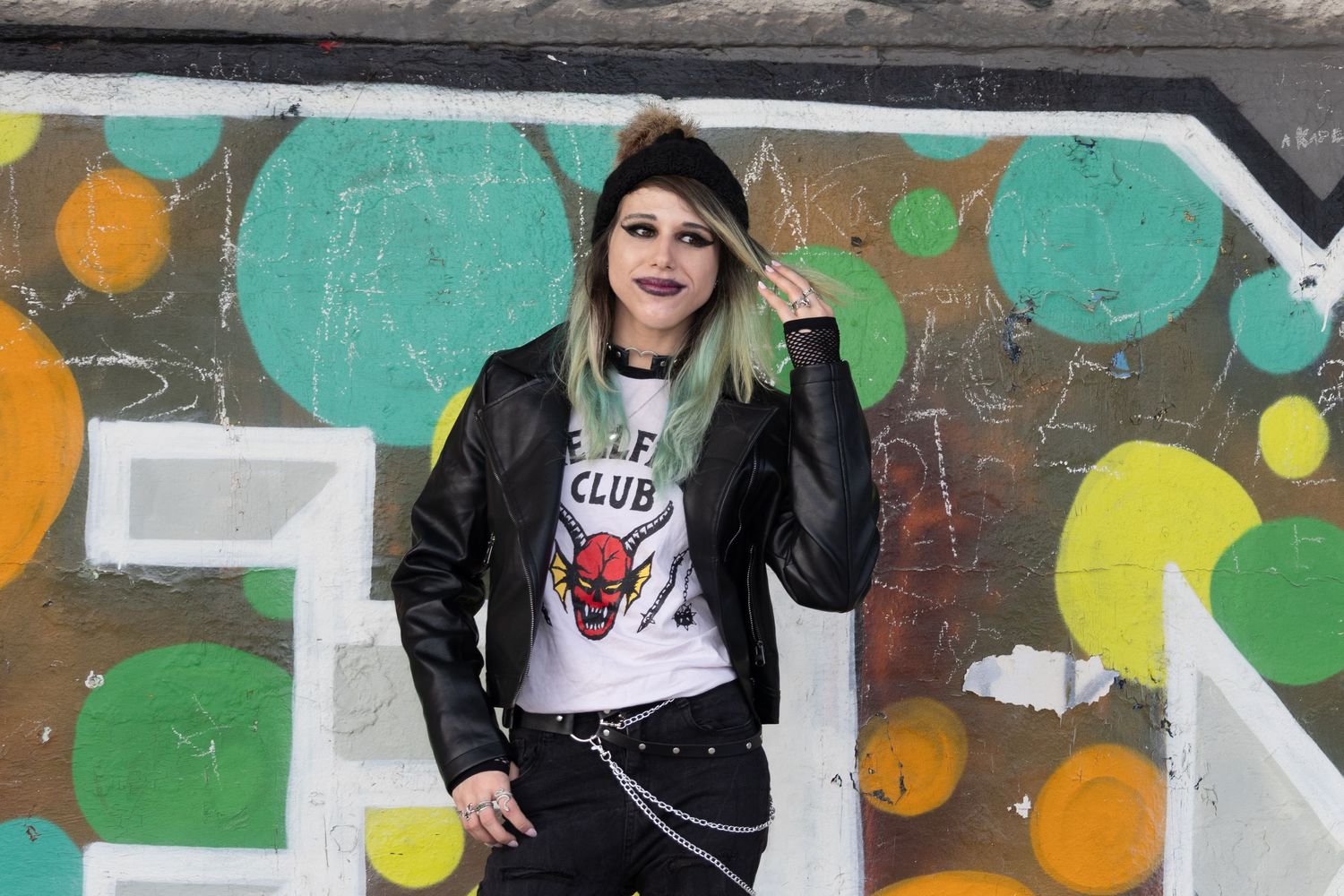 Young woman with green hair in leather jacket poses before colorful graffiti wall wearing Hellfire Club tee.