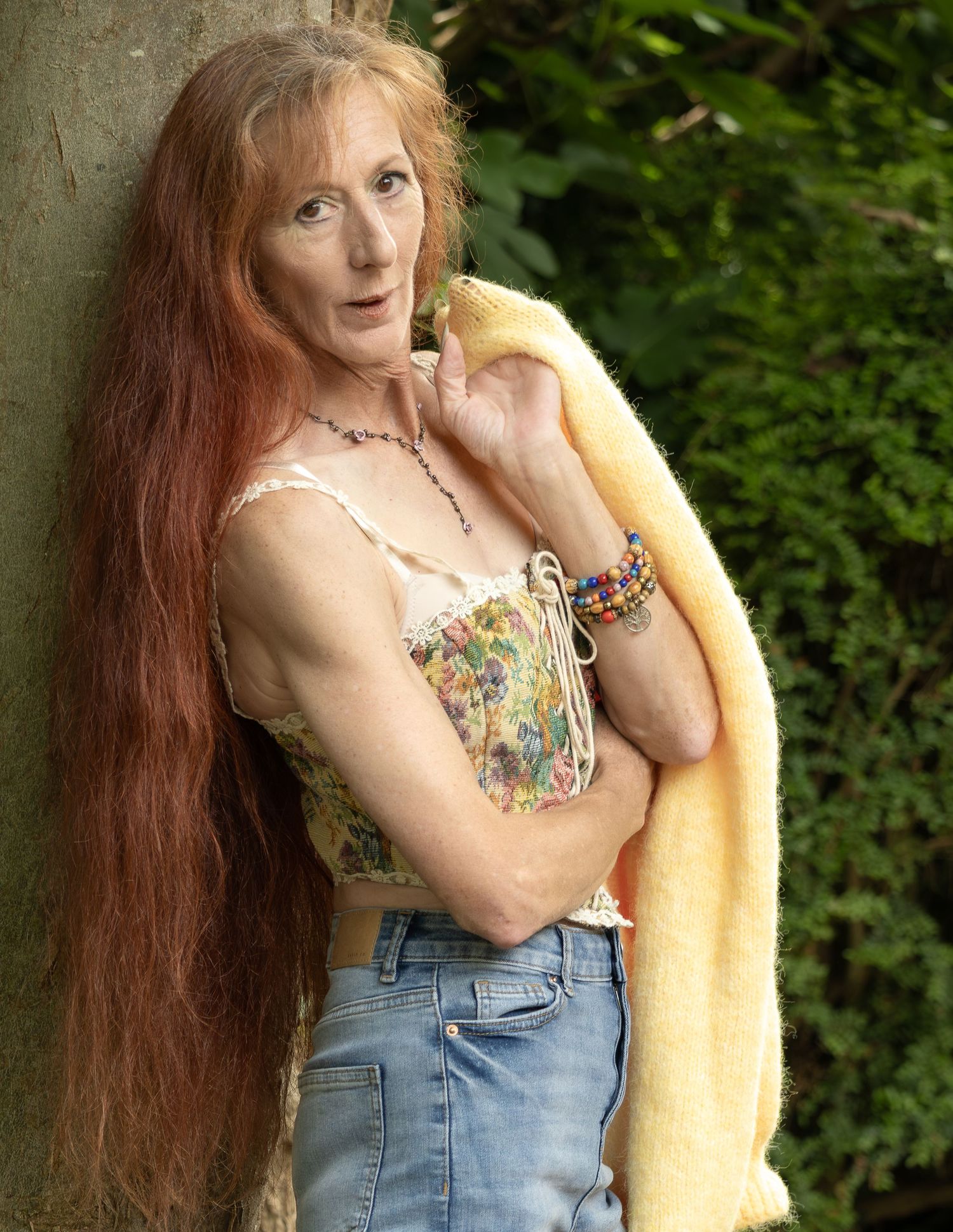 Older woman with long red hair in floral top, jeans, and yellow cardigan leaning against tree outdoors.