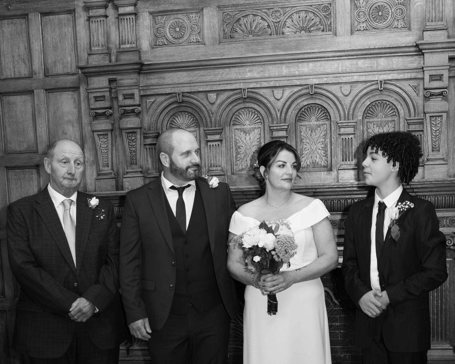 Black and white wedding portrait of bride, groom, and two family members before ornate wooden fireplace.