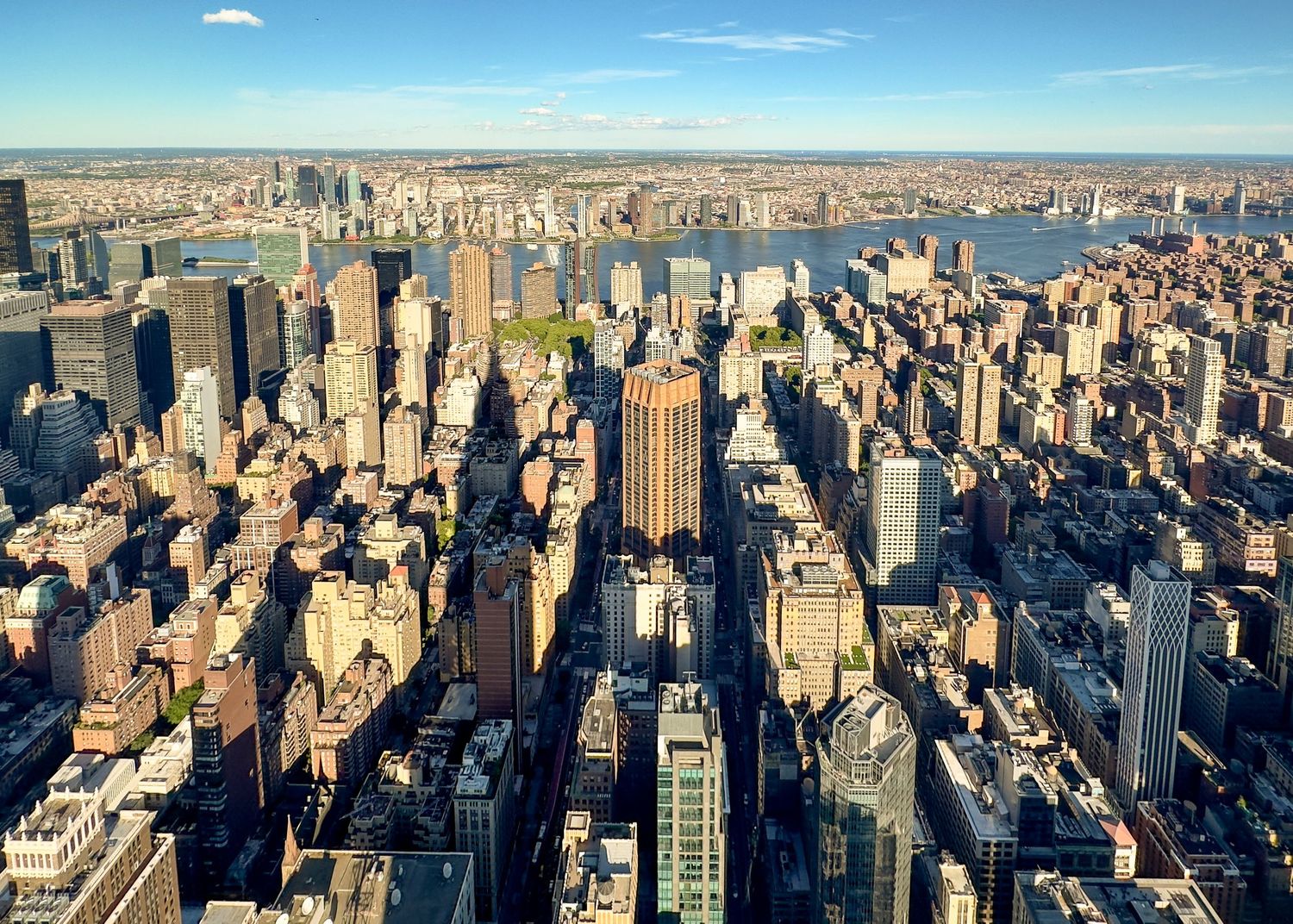 Aerial view of Manhattan skyline with dense skyscrapers, green parks, and the Hudson River under a clear blue sky.