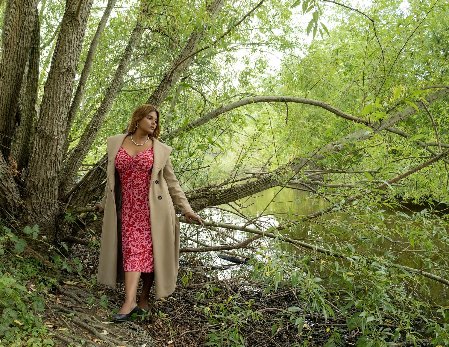 Woman in red lace dress and beige coat walking along a wooded creek path among tall trees.