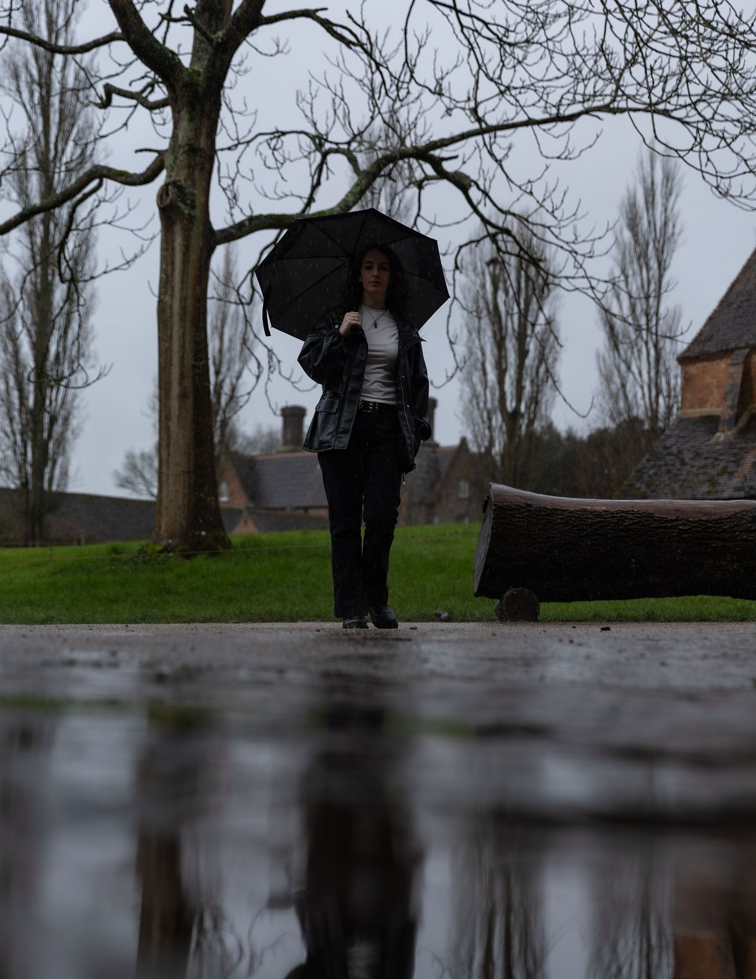 Woman walking with black umbrella in rainy park, reflected in puddle on overcast day.