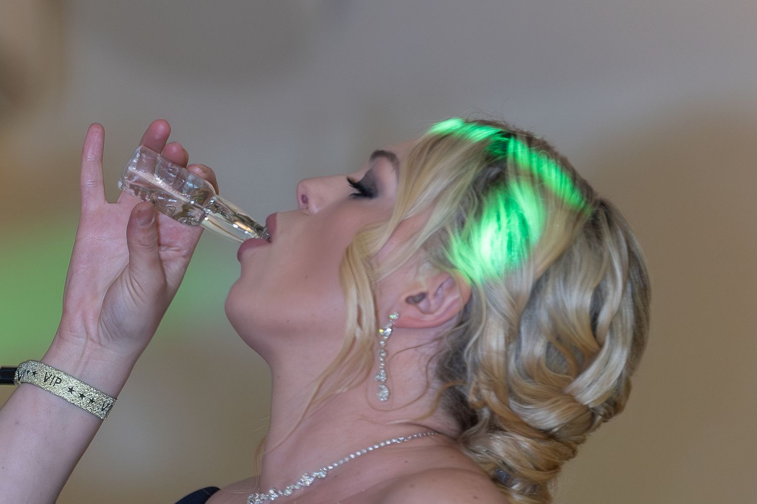 Blonde woman drinking from a small glass bottle at a party with green lights reflecting in her hair.