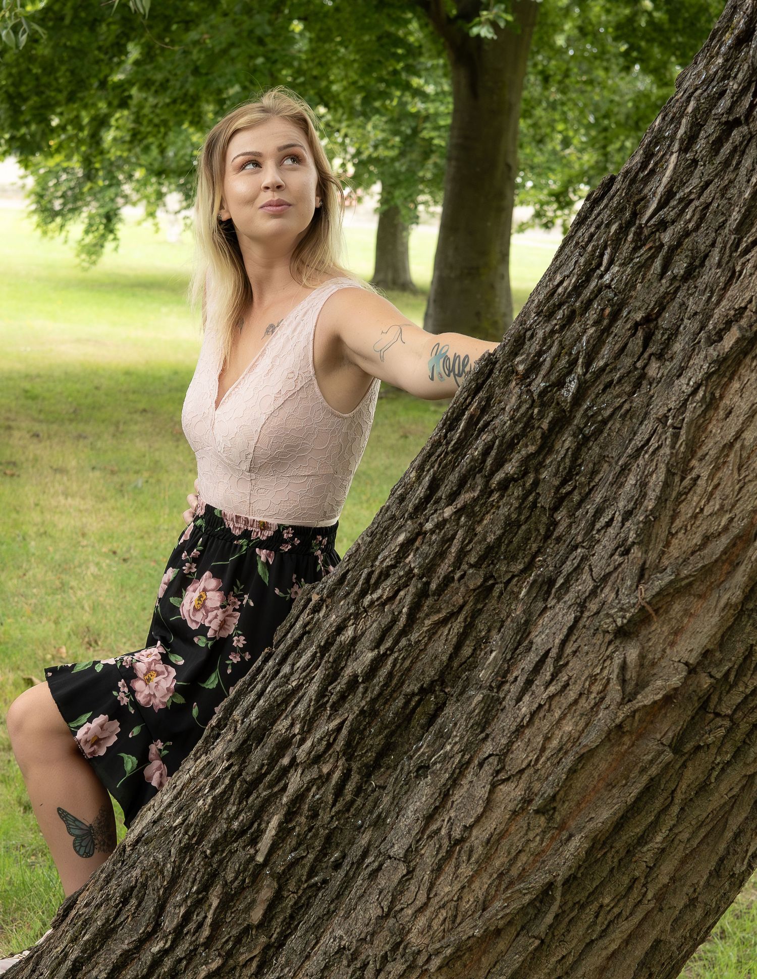 A tattooed blonde woman in a floral skirt leans against a large tree trunk in a lush green park.