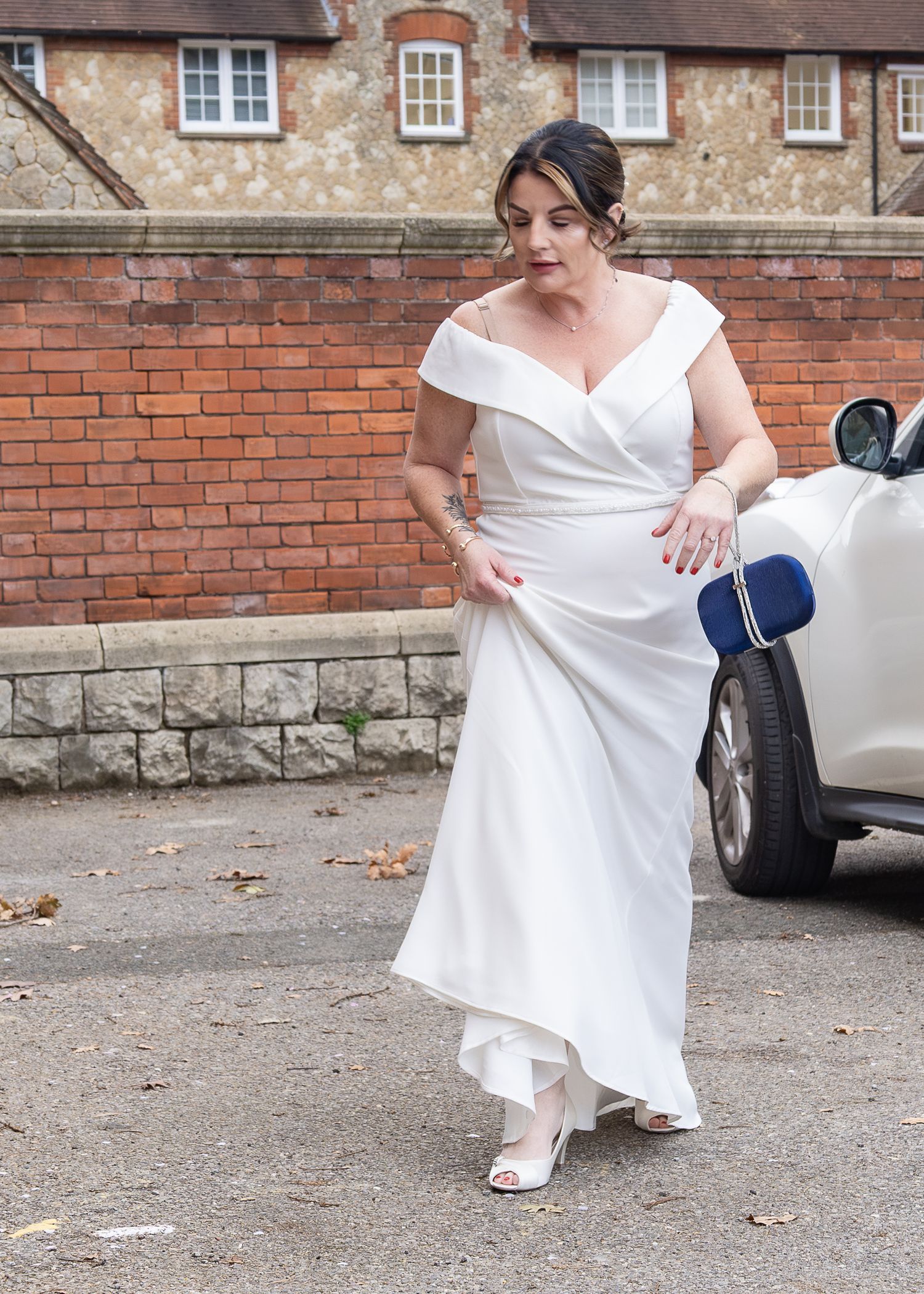 Woman in white off-shoulder wedding gown holding a blue clutch bag, walking near a brick building.