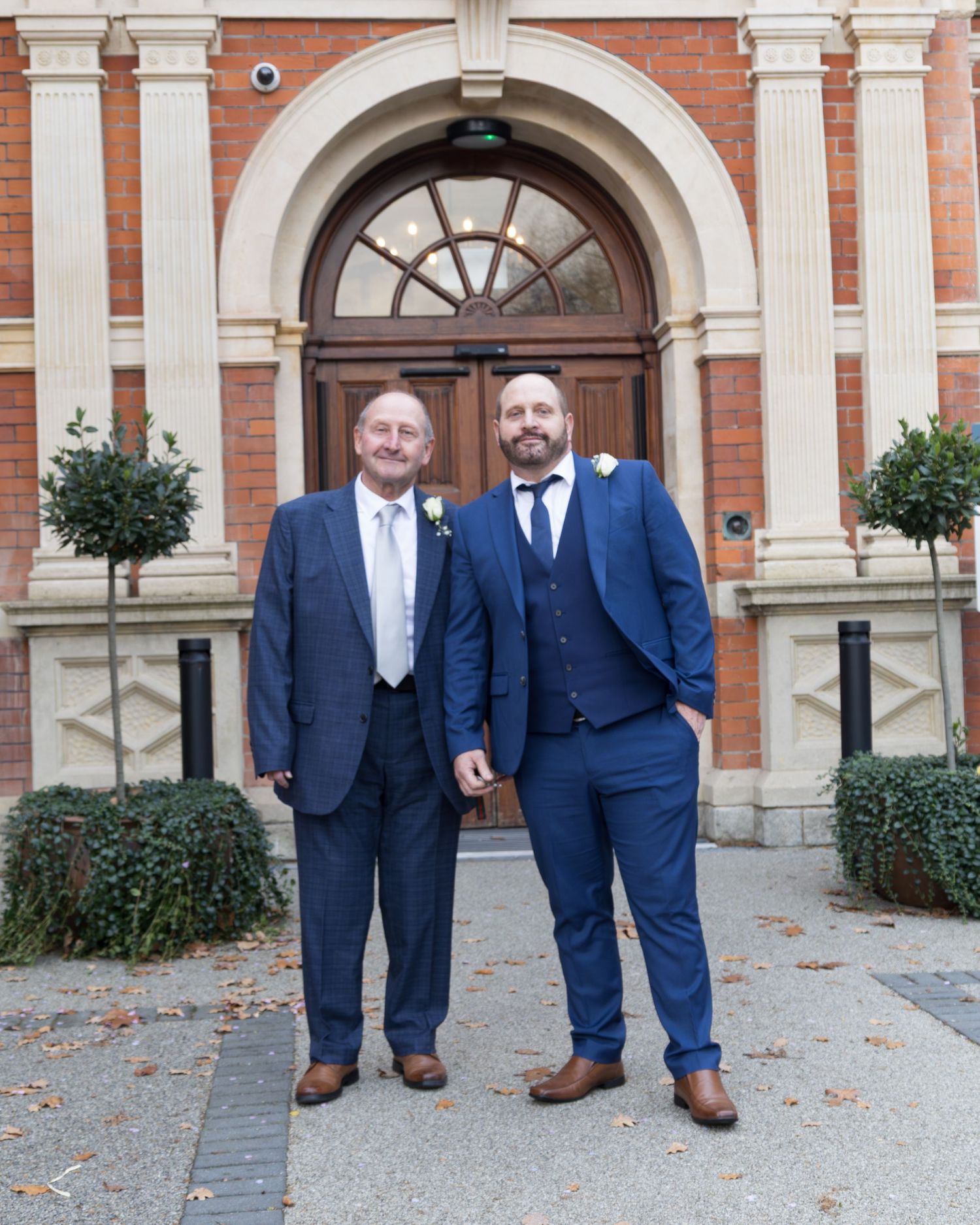 Two men in navy blue suits with boutonnieres pose outside a red brick building with ornate columns.