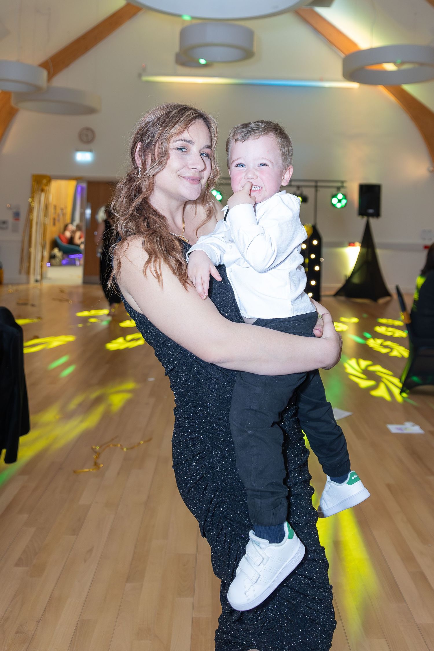 Woman in black dress holding a toddler boy in white outfit on a dance floor with colorful lights.