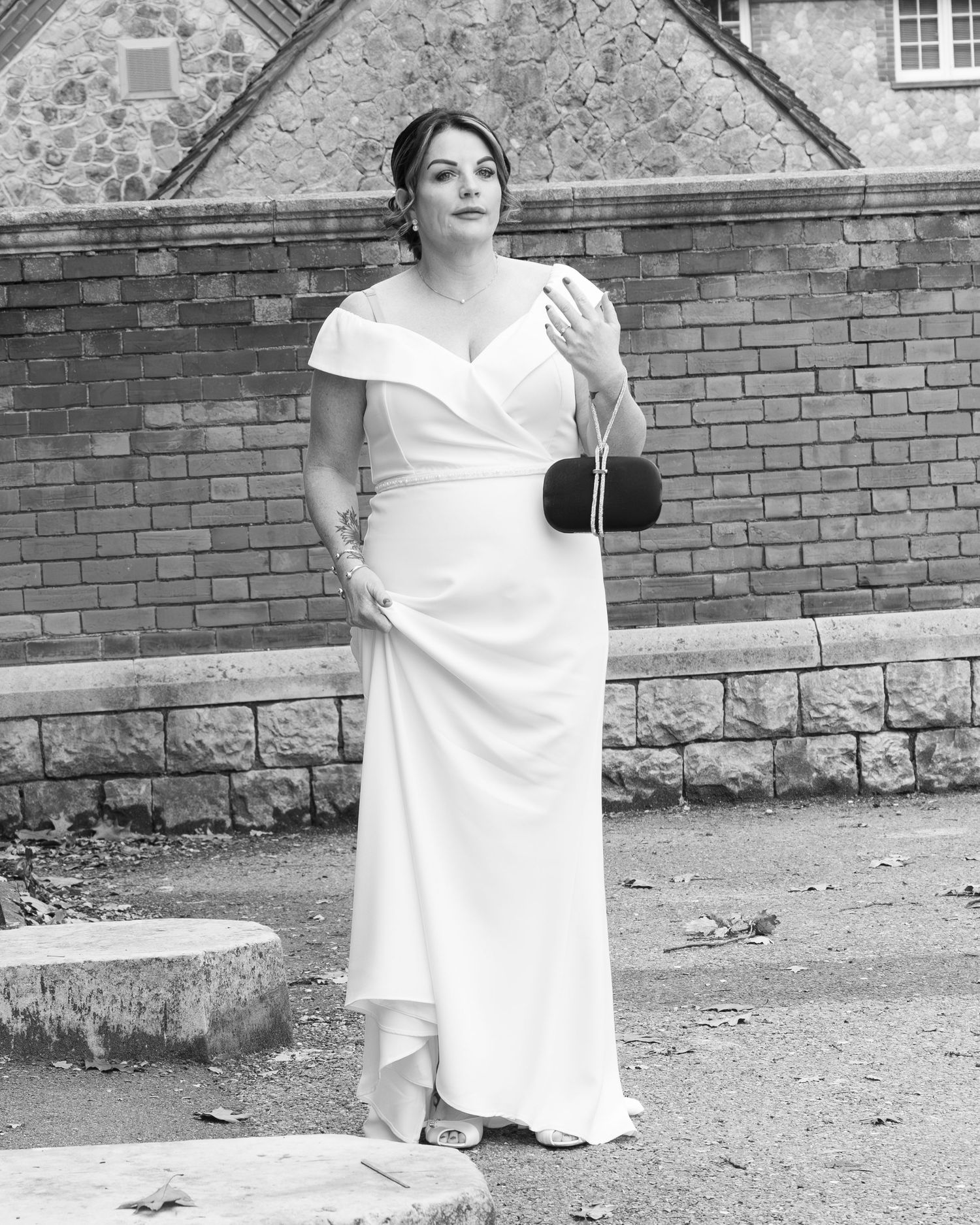 Bride in elegant white off-shoulder gown holds black clutch purse outside stone building.