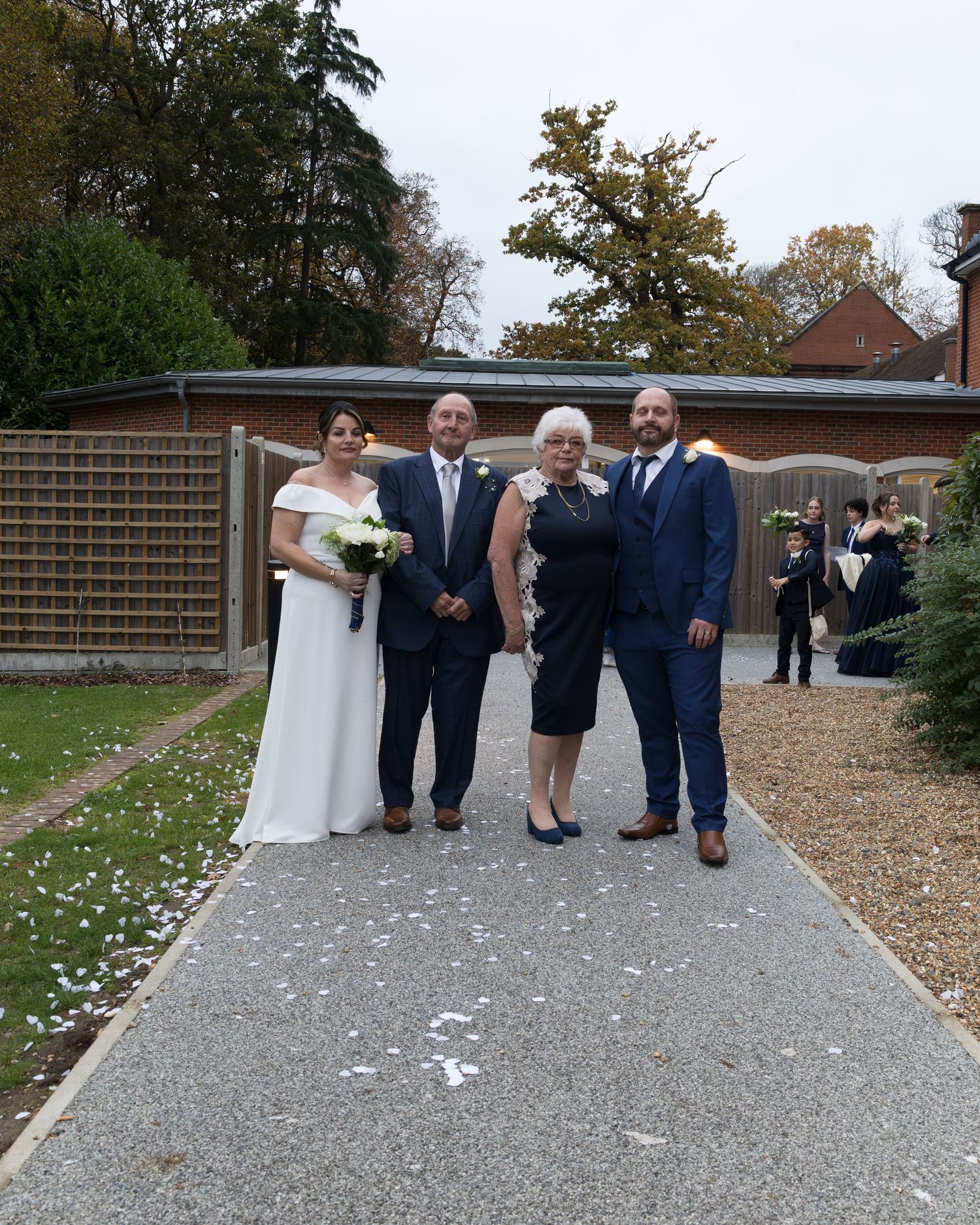 Wedding party of four poses on gravel path outside venue, bride holding bouquet.