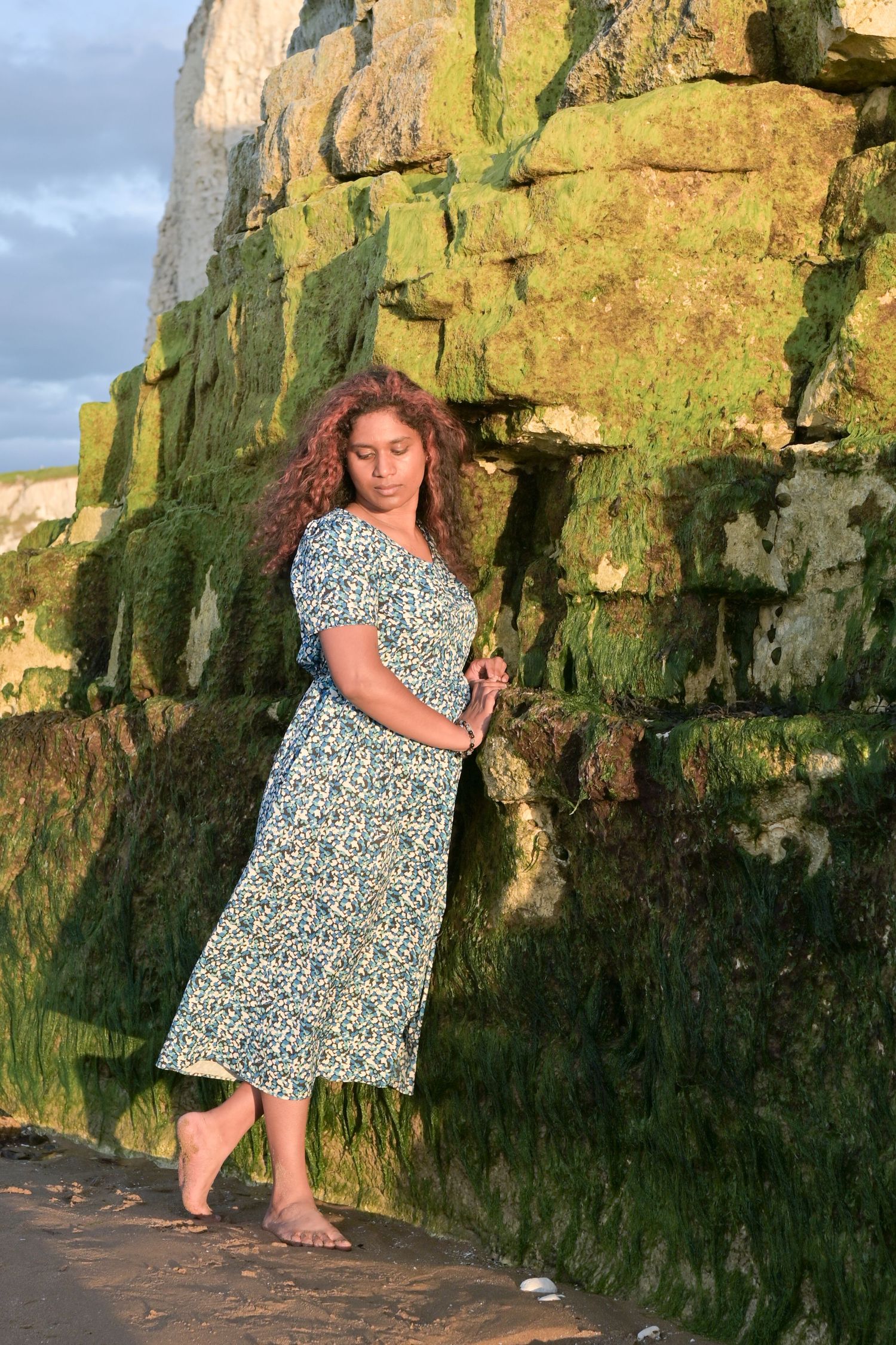 Woman in floral dress posing against moss-covered rocks at sunset near the ocean.
