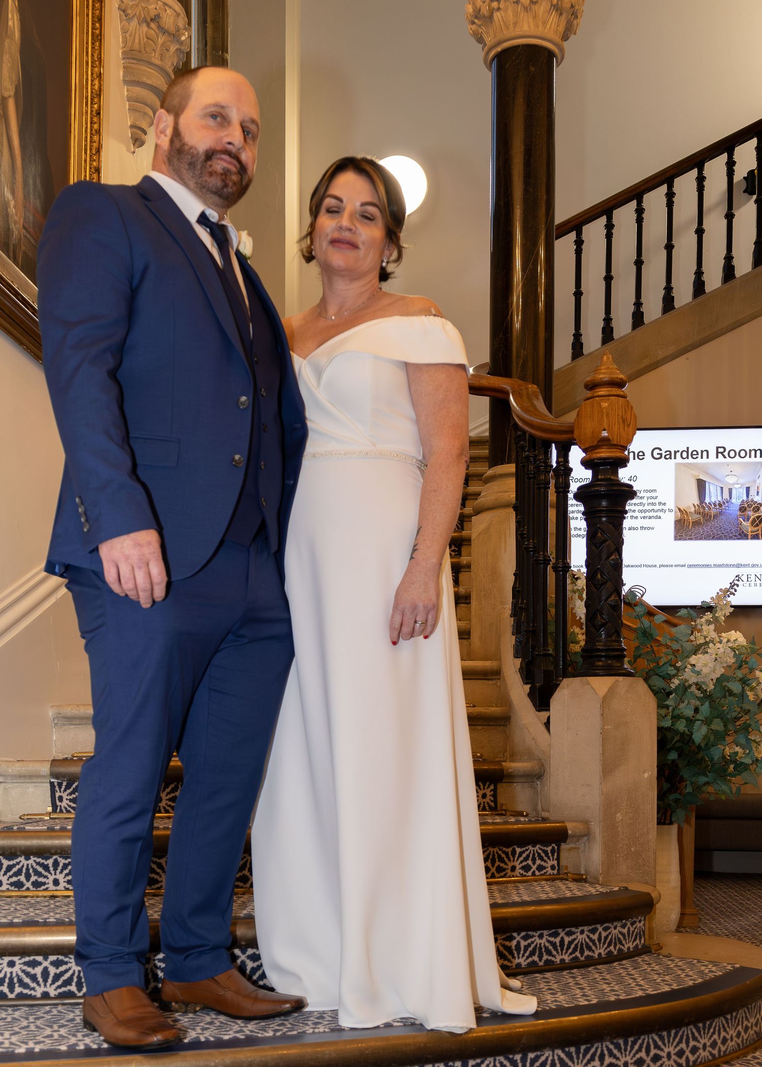 Bride in white off-shoulder gown and groom in navy suit pose elegantly on ornate staircase at wedding venue.