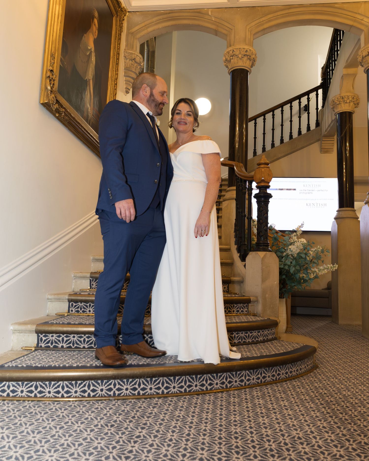 Bride in white off-shoulder gown and groom in navy suit posing on ornate tiled staircase at wedding.