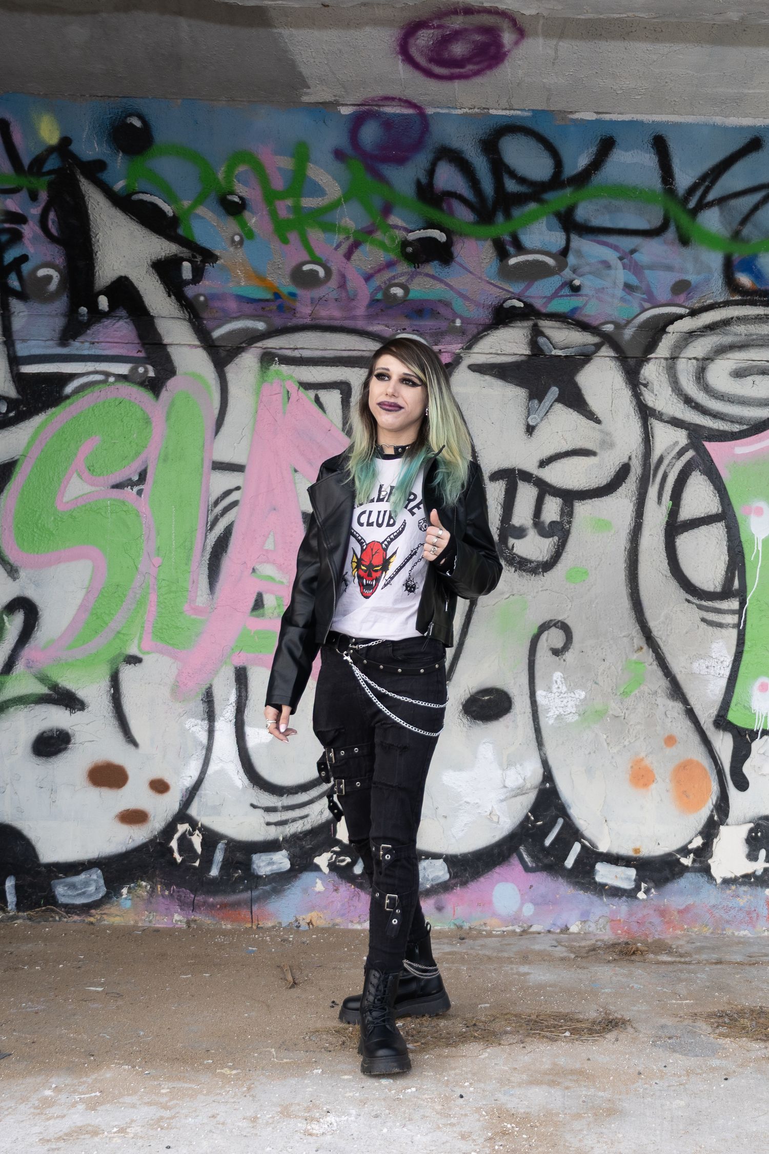 Young woman in punk style outfit with green hair poses before colorful graffiti wall in urban setting.