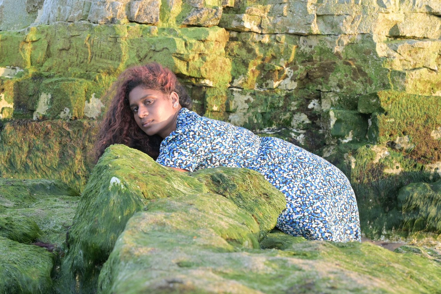Woman in blue floral dress posing on moss-covered rocks by a stone wall outdoors.
