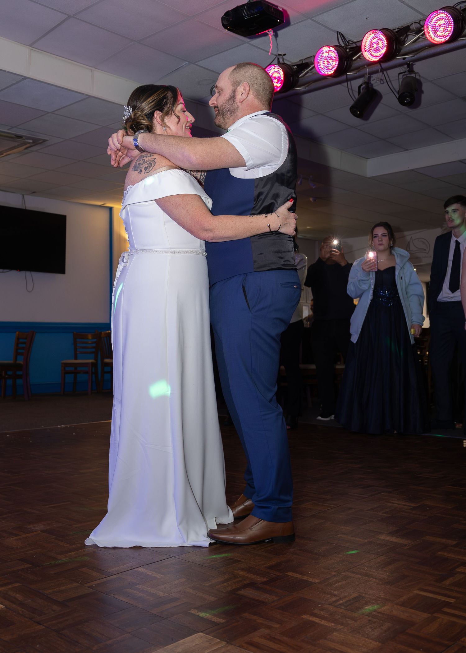 Bride and groom share their first dance at wedding reception, embraced on dance floor with colorful stage lights above.