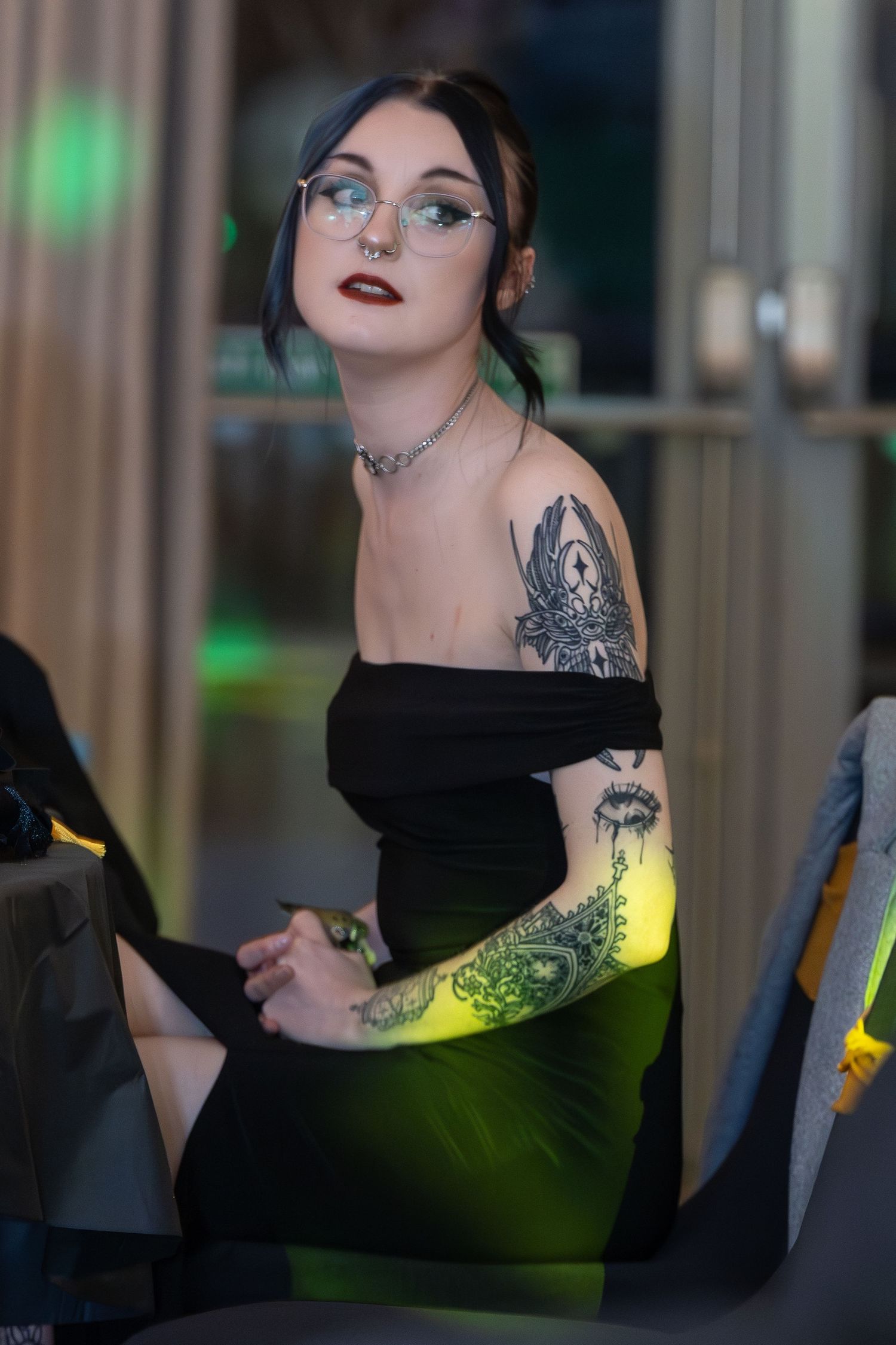 Young woman in black off-shoulder dress with tattoos and glasses posing at an event with green lighting.