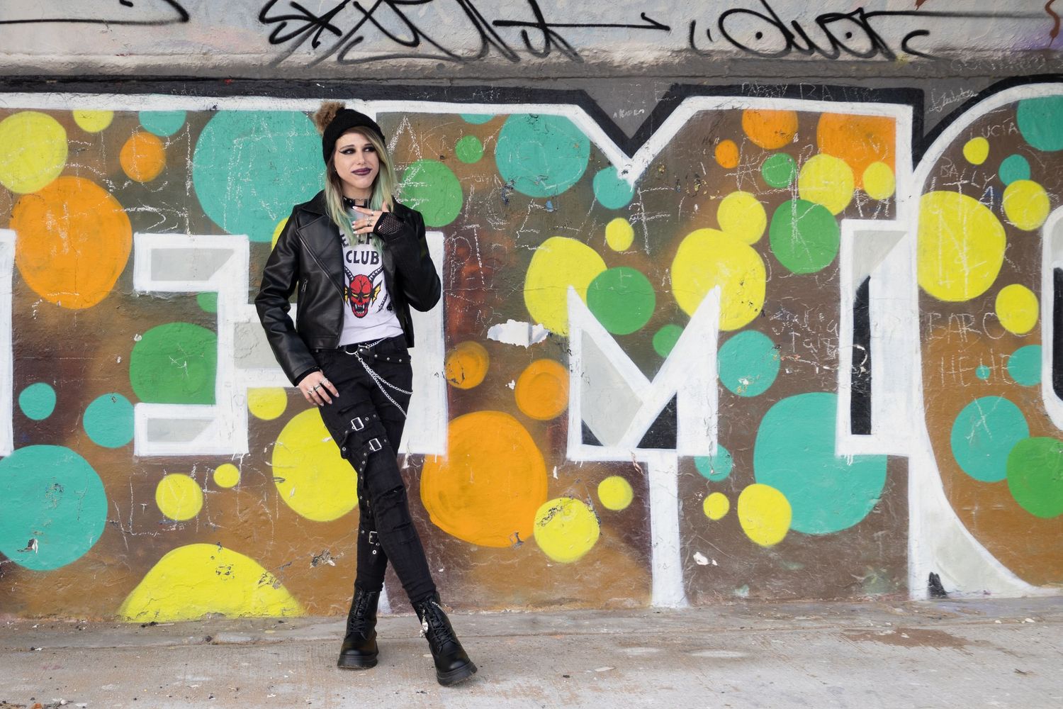 A punk-style person poses in front of colorful graffiti wall with EMO text and polka dot design outdoors.