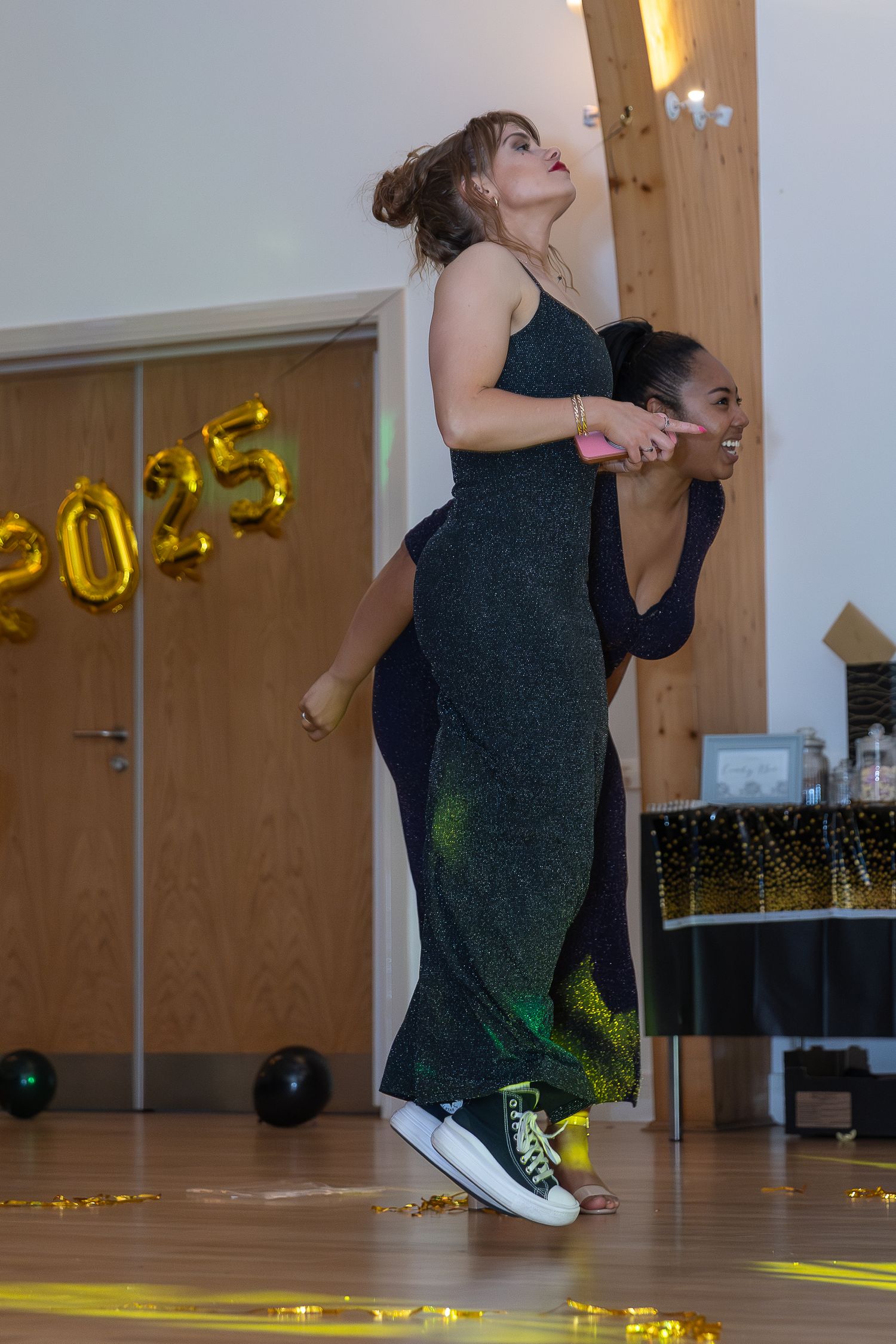 Two young women in formal gowns dancing at a 2025 prom celebration on a decorated gymnasium floor.