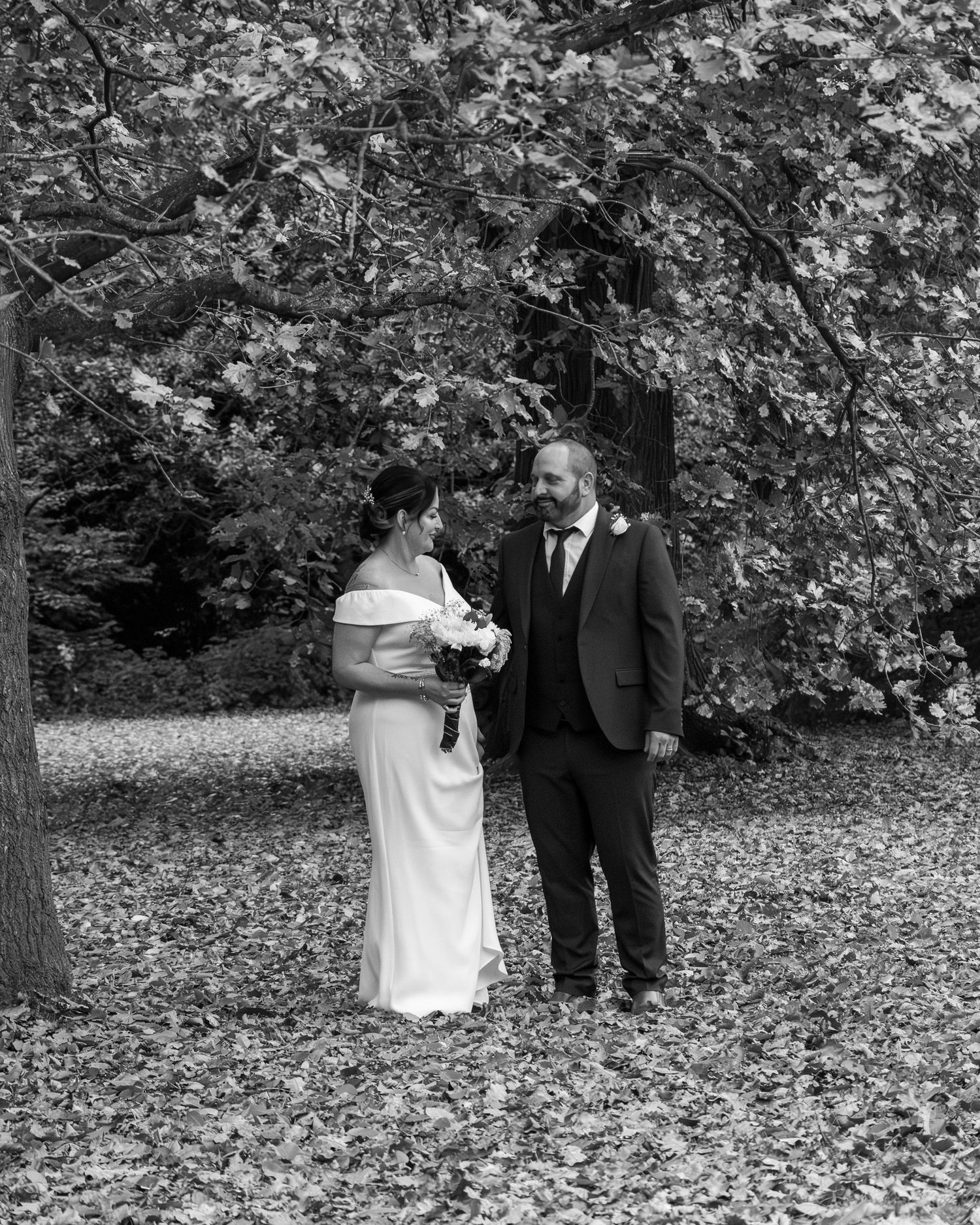 Black and white photo of a bride and groom standing among fallen leaves beneath a large tree.