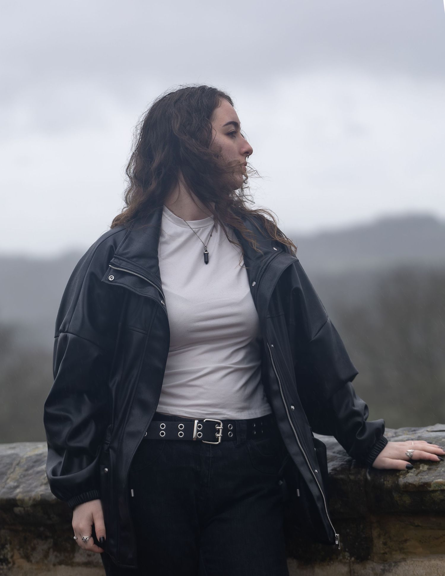Young woman in black leather jacket and white top standing outdoors on a cloudy misty day.