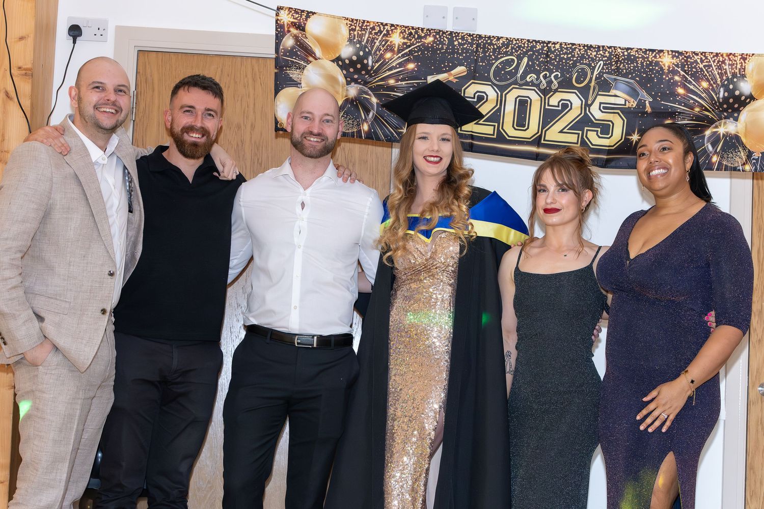 Group of six people posing at a Class of 2025 graduation celebration, with one woman in cap and gown.